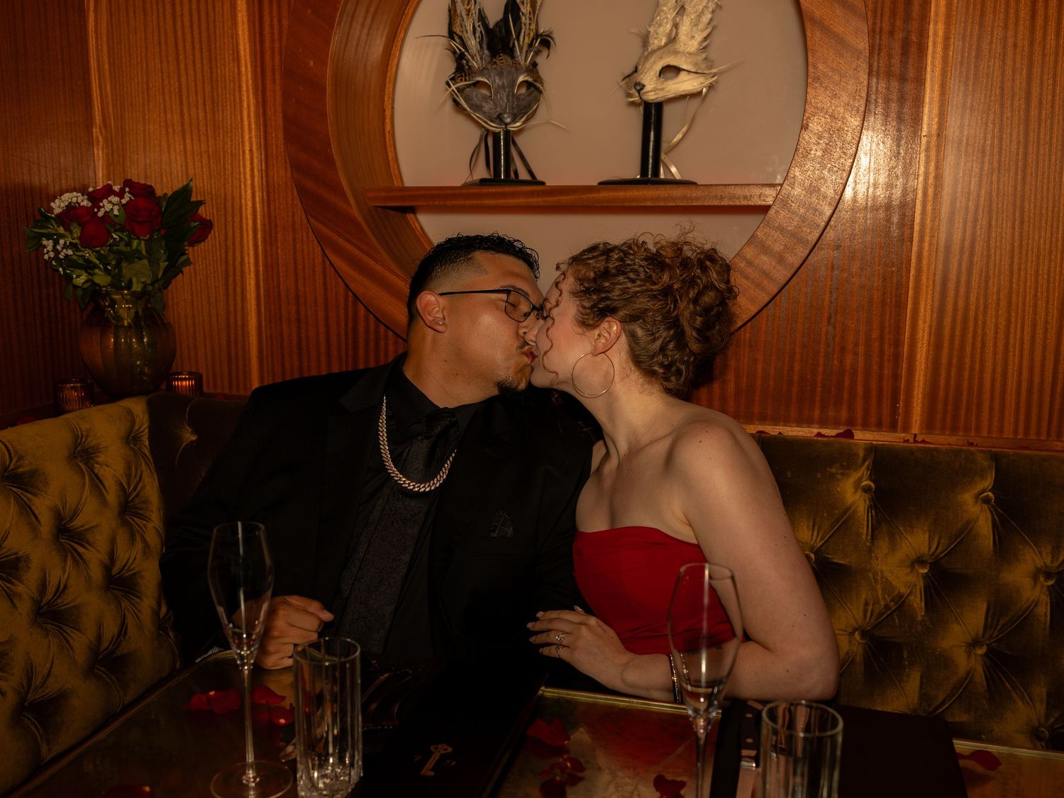 Couple sharing a romantic kiss at an elegant restaurant booth with champagne glasses and roses.