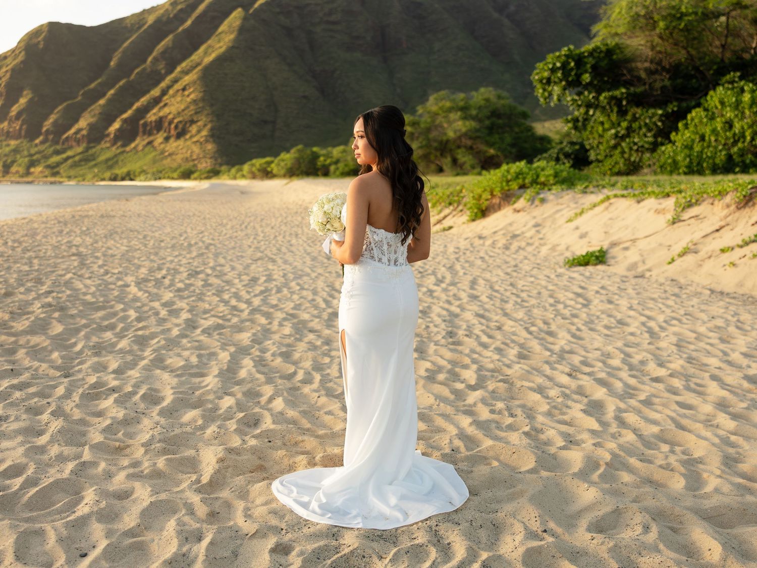 Bride in white lace gown holding bouquet on sandy beach with lush green mountains in background.