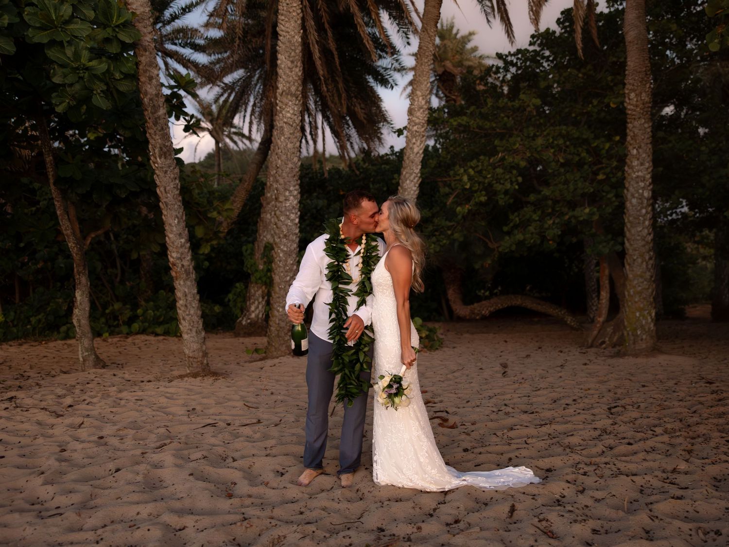 Couple sharing a kiss on a sandy beach surrounded by palm trees during their Hawaiian wedding.