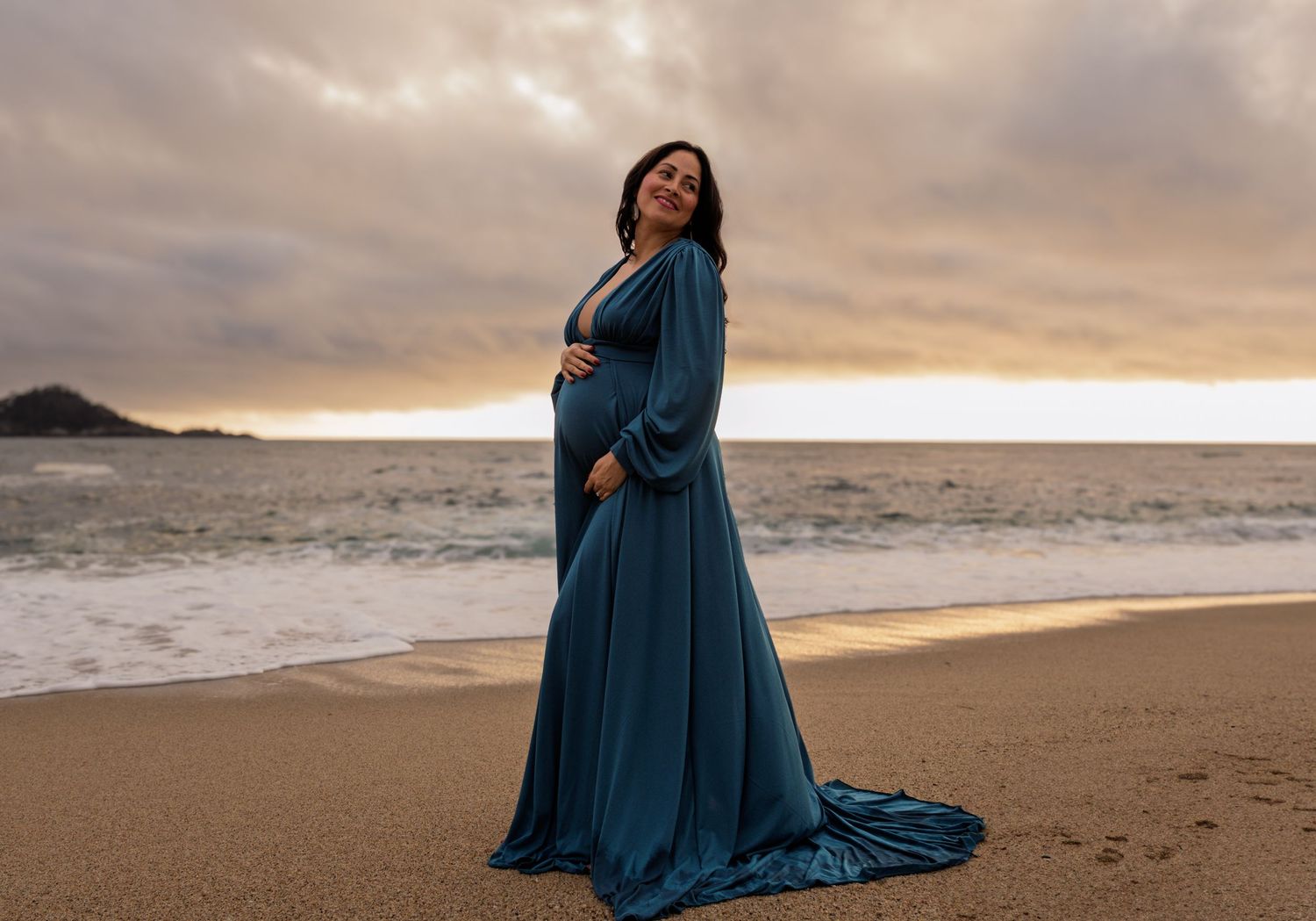 Pregnant woman in flowing teal gown posing on beach at dramatic sunset.