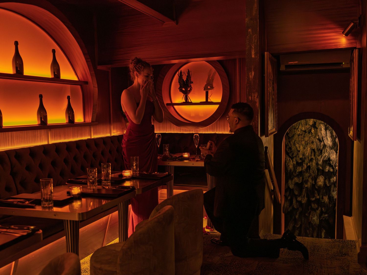 Man proposing on one knee to woman in dimly lit romantic restaurant with warm orange ambient lighting.