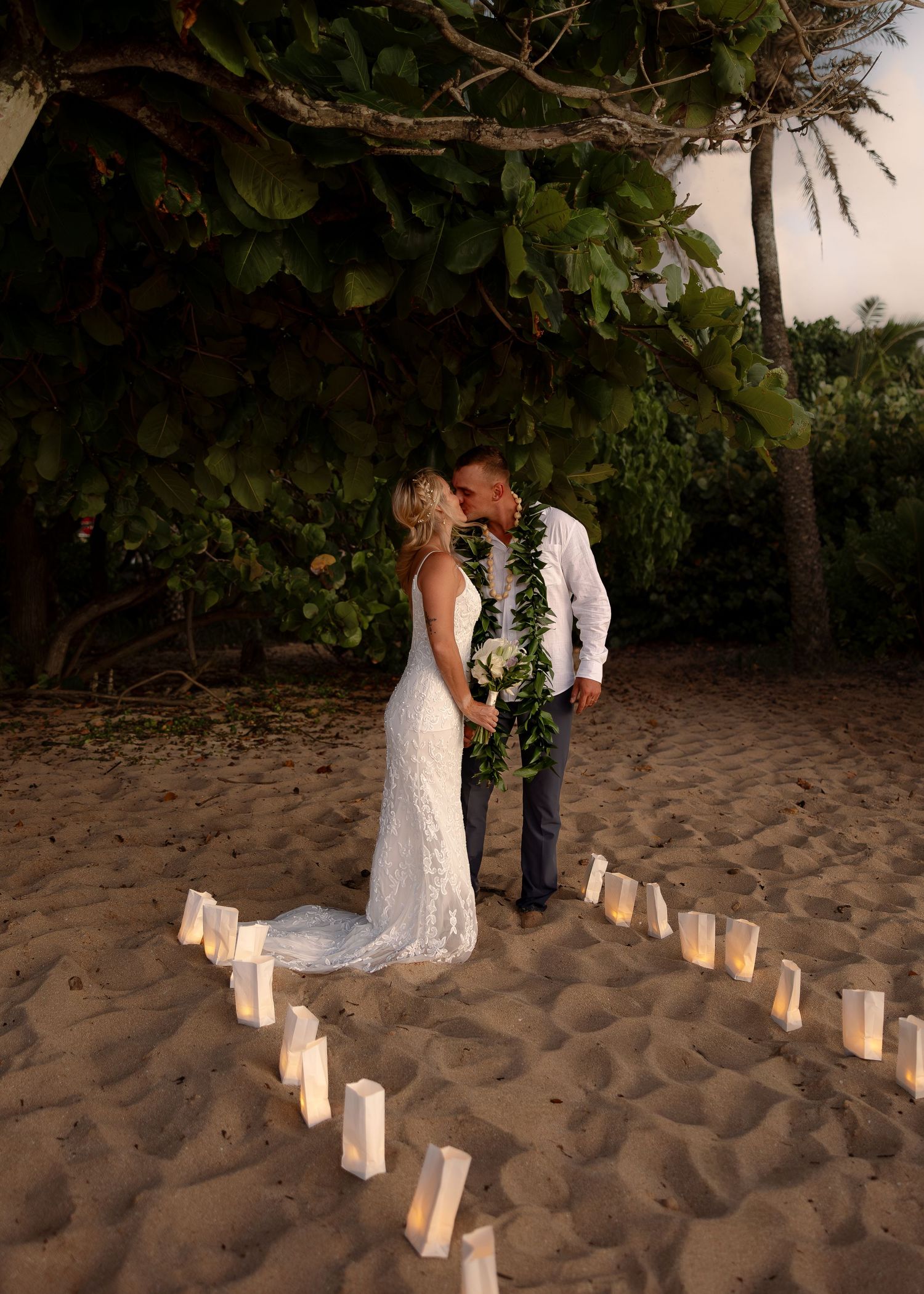 Couple sharing a kiss at a romantic beach wedding ceremony surrounded by glowing candle luminaries at sunset in Hawaii.