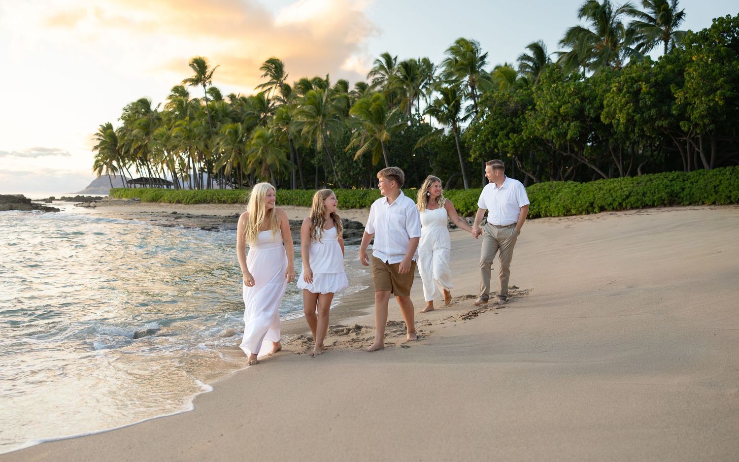 Family of five walking on a tropical beach at sunset, wearing white outfits with palm trees in the background.