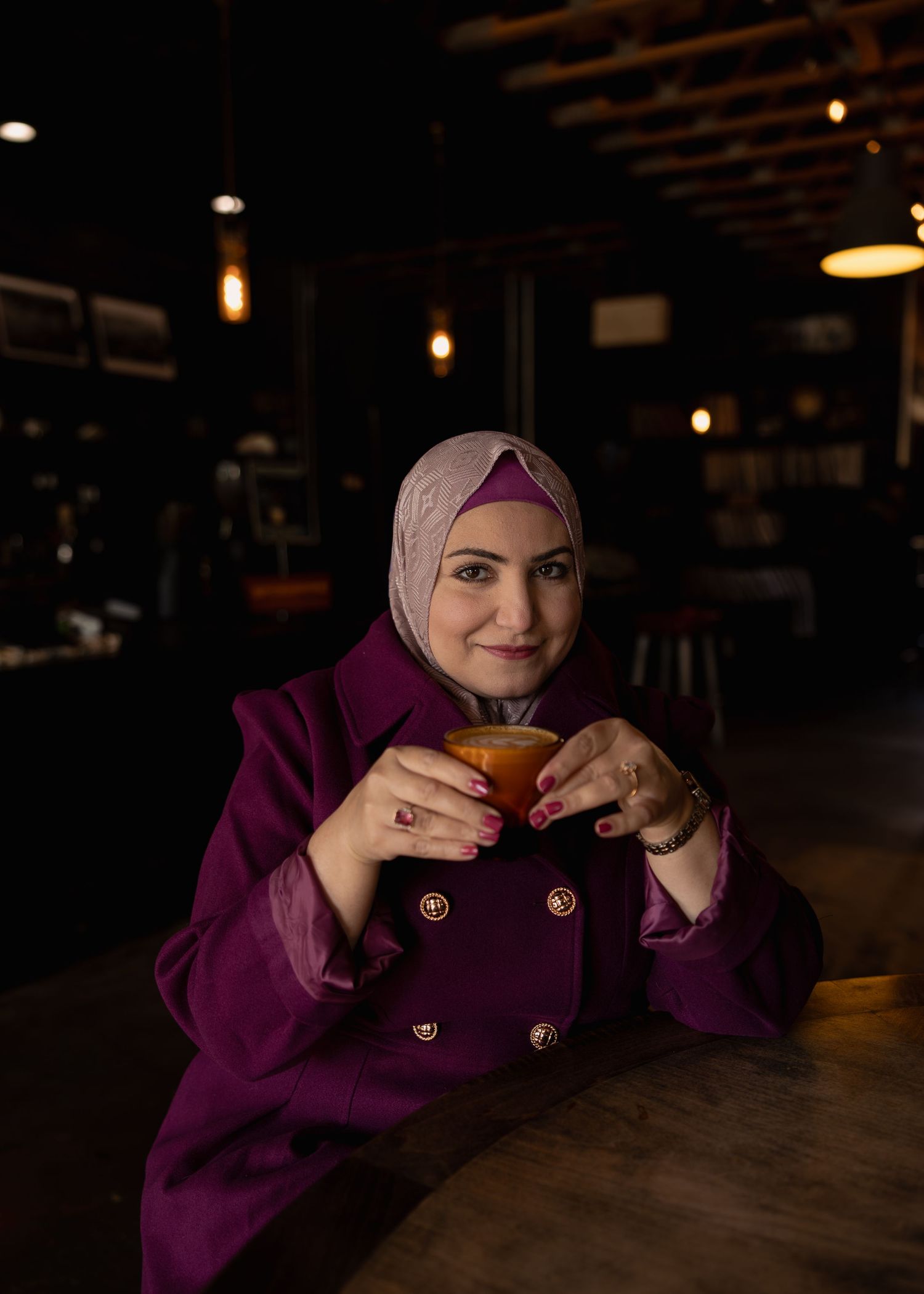 Woman in purple coat and hijab holding a warm drink at a cozy cafe.
