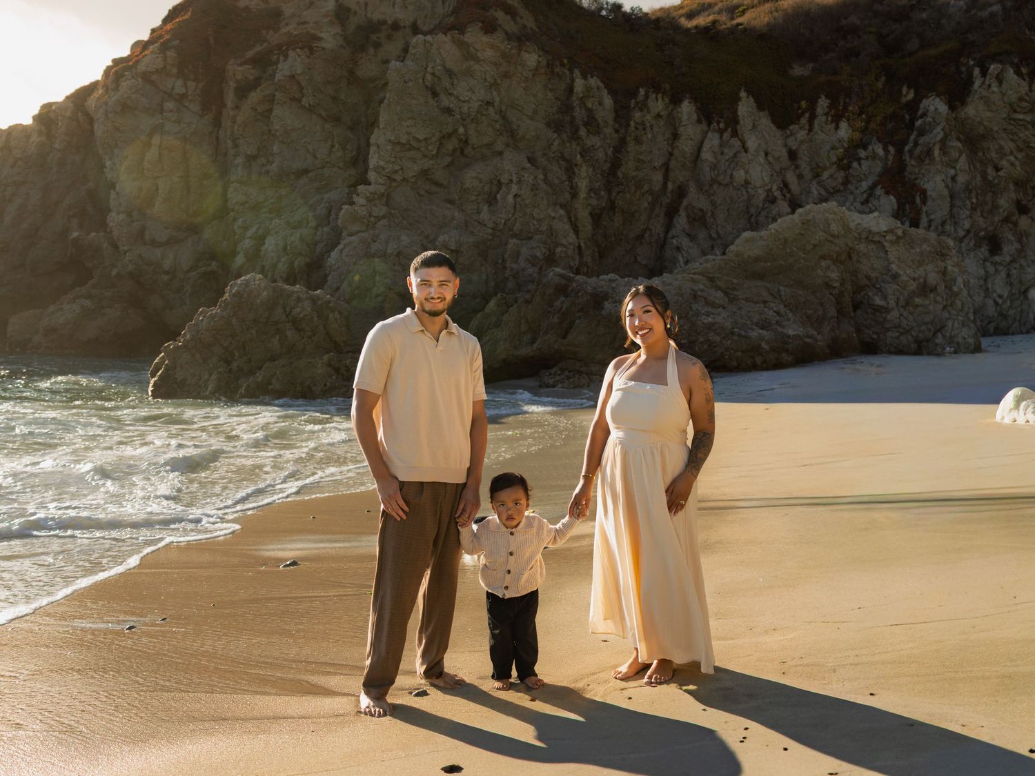 Young family of three holding hands on sunlit beach with dramatic cliffs and ocean waves behind them.