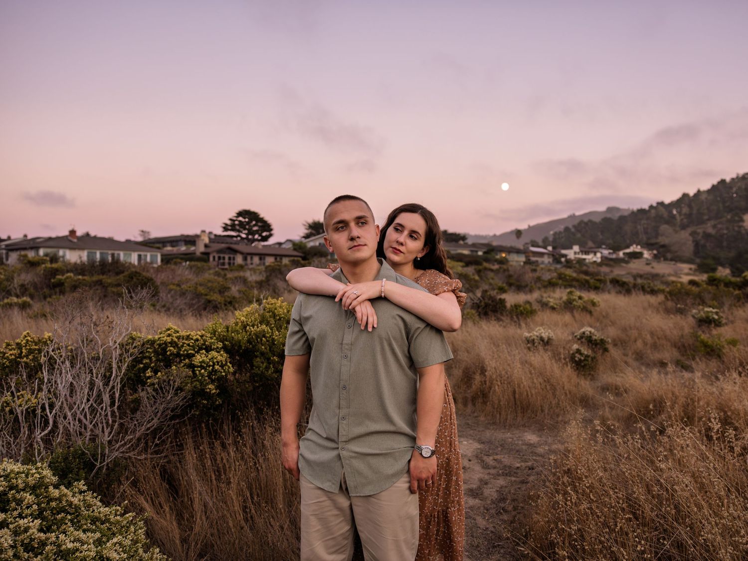 Couple posing at dusk in open field with full moon rising over hills and homes in background.