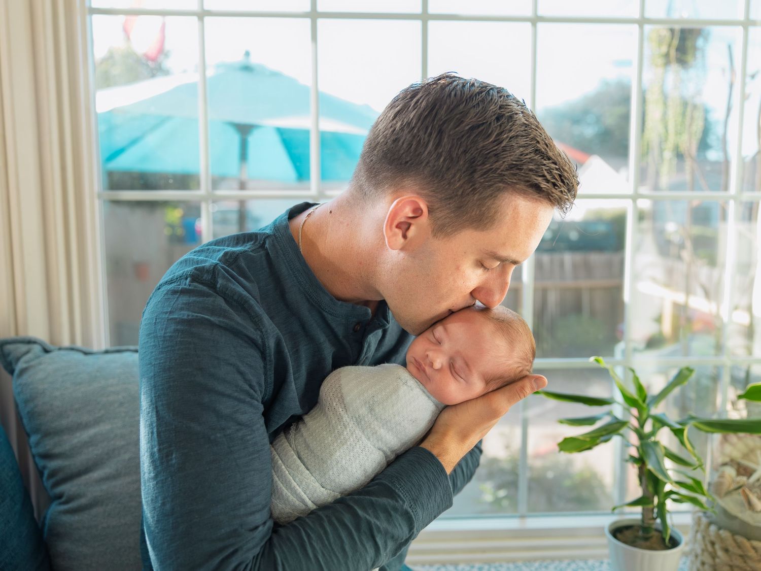 Father tenderly kissing his newborn baby wrapped in white swaddle blanket by a bright sunny window.