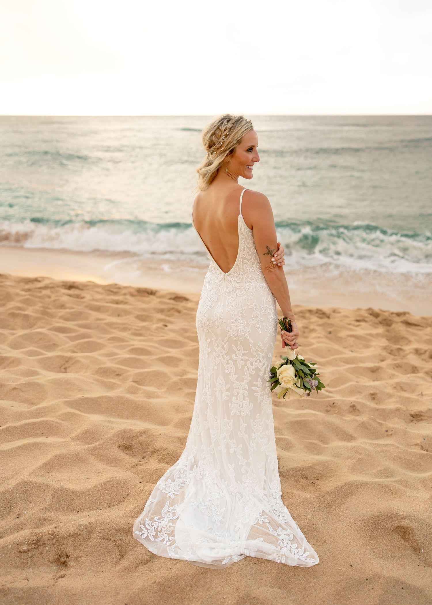 Bride in a backless lace wedding gown holding a bouquet, standing on a sandy beach with ocean waves behind her.