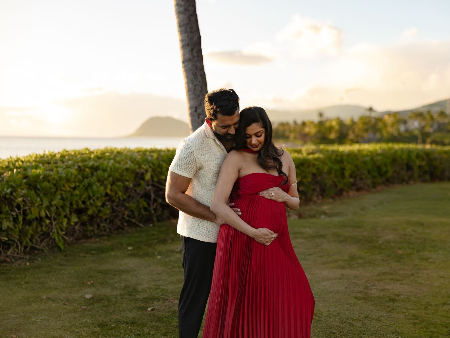 Couple in maternity photoshoot at sunset, woman in red pleated gown cradling baby bump, man in white shirt embracing her.