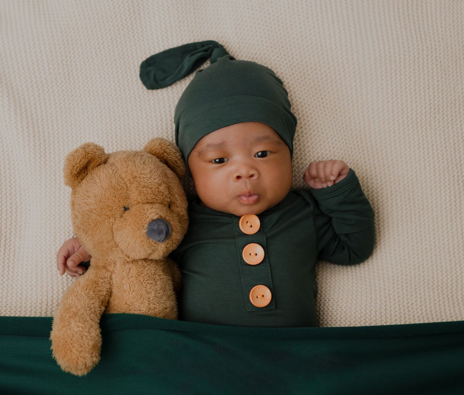 Newborn baby in green knot hat and button romper lying next to a brown teddy bear on a cream blanket.