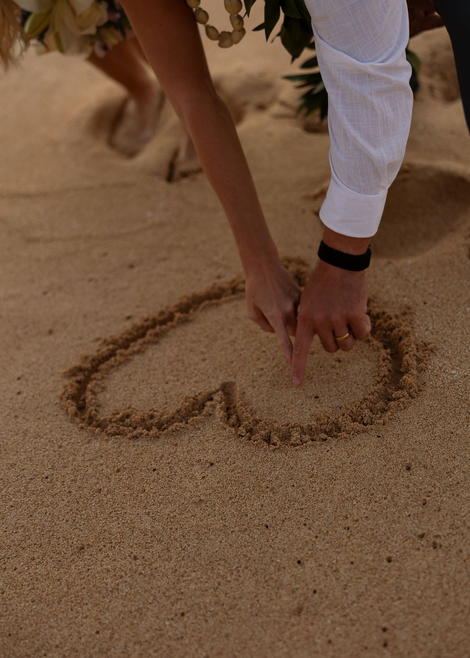 Couple drawing a heart shape in sandy beach, wearing wedding rings and holding bouquet of flowers.