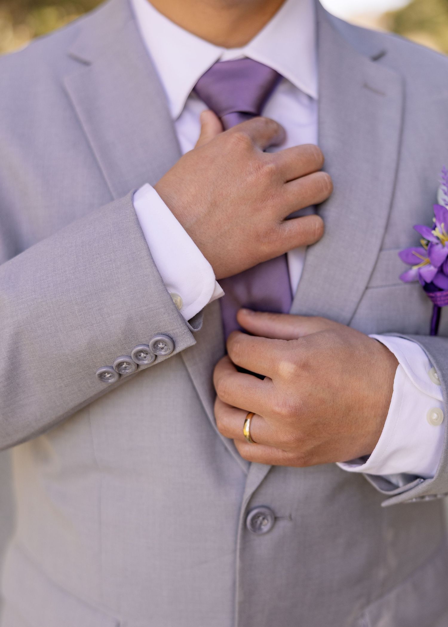 Groom in gray suit adjusting purple tie with gold wedding band, purple boutonniere visible on lapel.