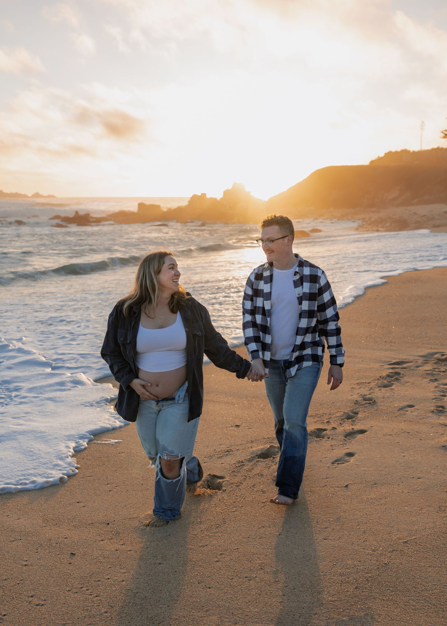 Couple holding hands walking on a sandy beach at golden sunset, woman showing baby bump in white crop top.