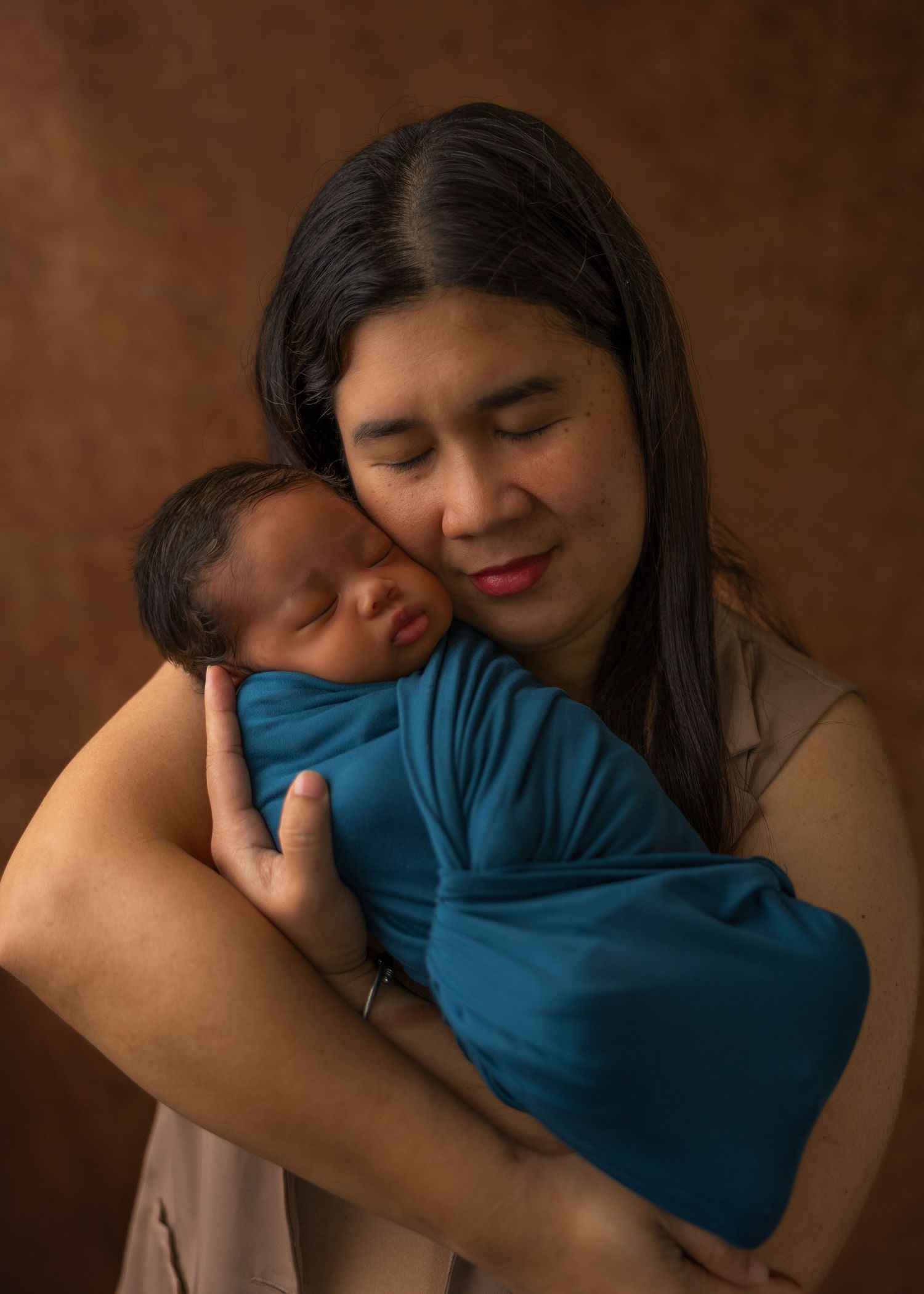 A mother tenderly holds her swaddled newborn baby wrapped in teal against a warm brown studio backdrop.