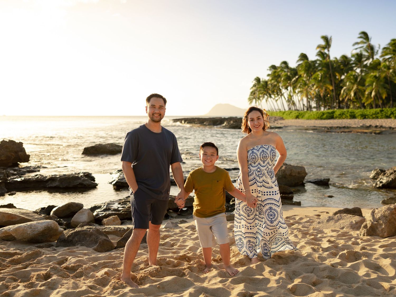 Family of three posing on a sandy tropical beach at sunset with palm trees and ocean in the background.
