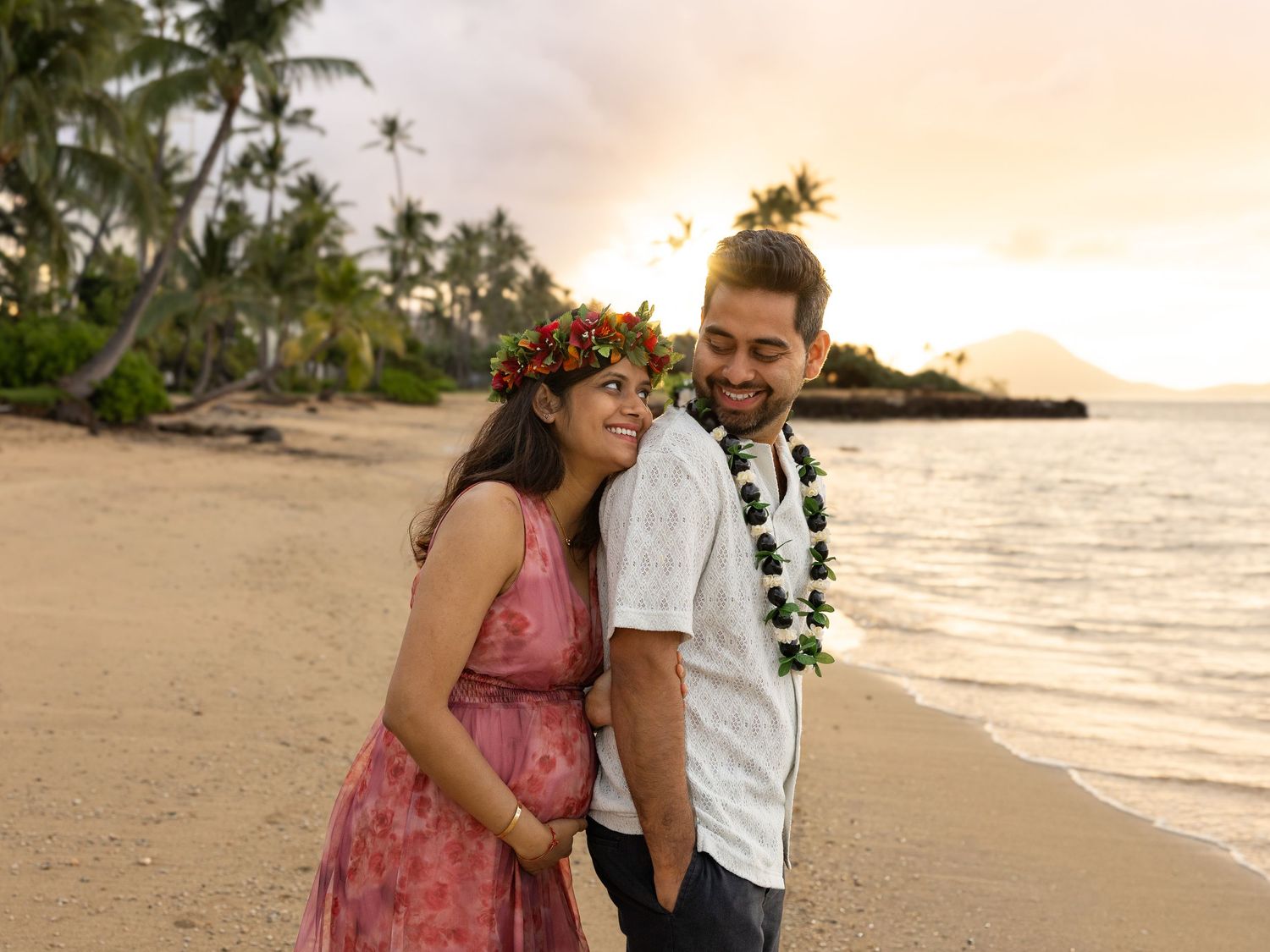 Pregnant couple in Hawaiian leis poses on a sandy beach at sunset with palm trees and ocean in the background.