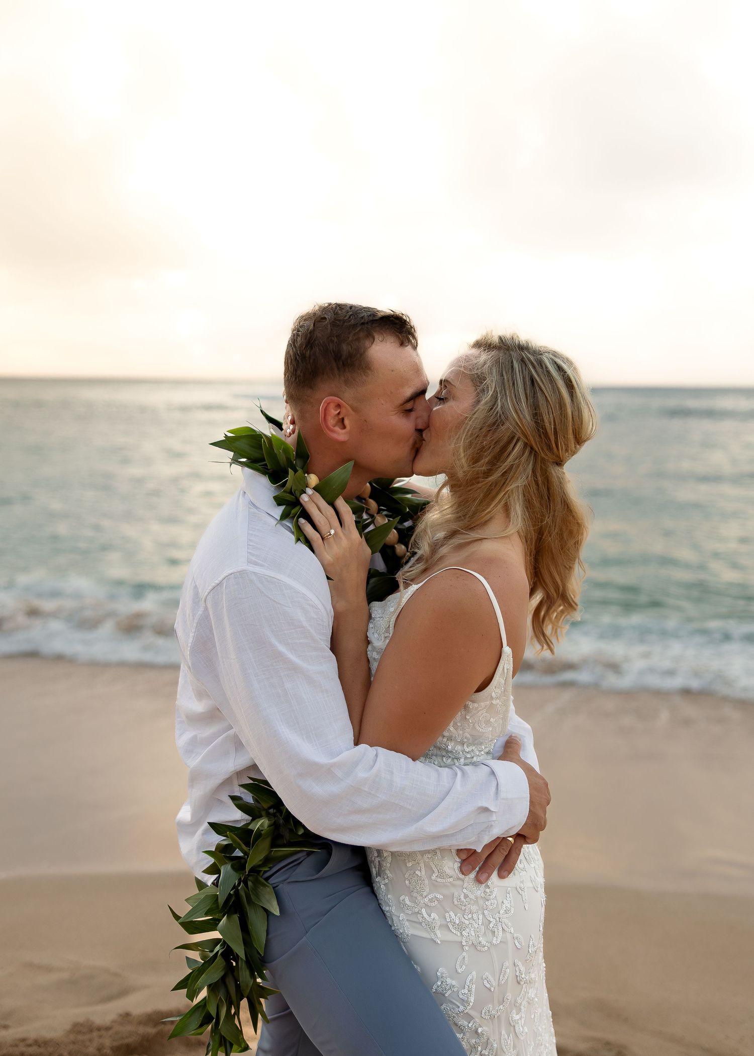 Couple sharing a romantic kiss on a Hawaiian beach at sunset, groom wearing a traditional lei and bride in a lace wedding gown.