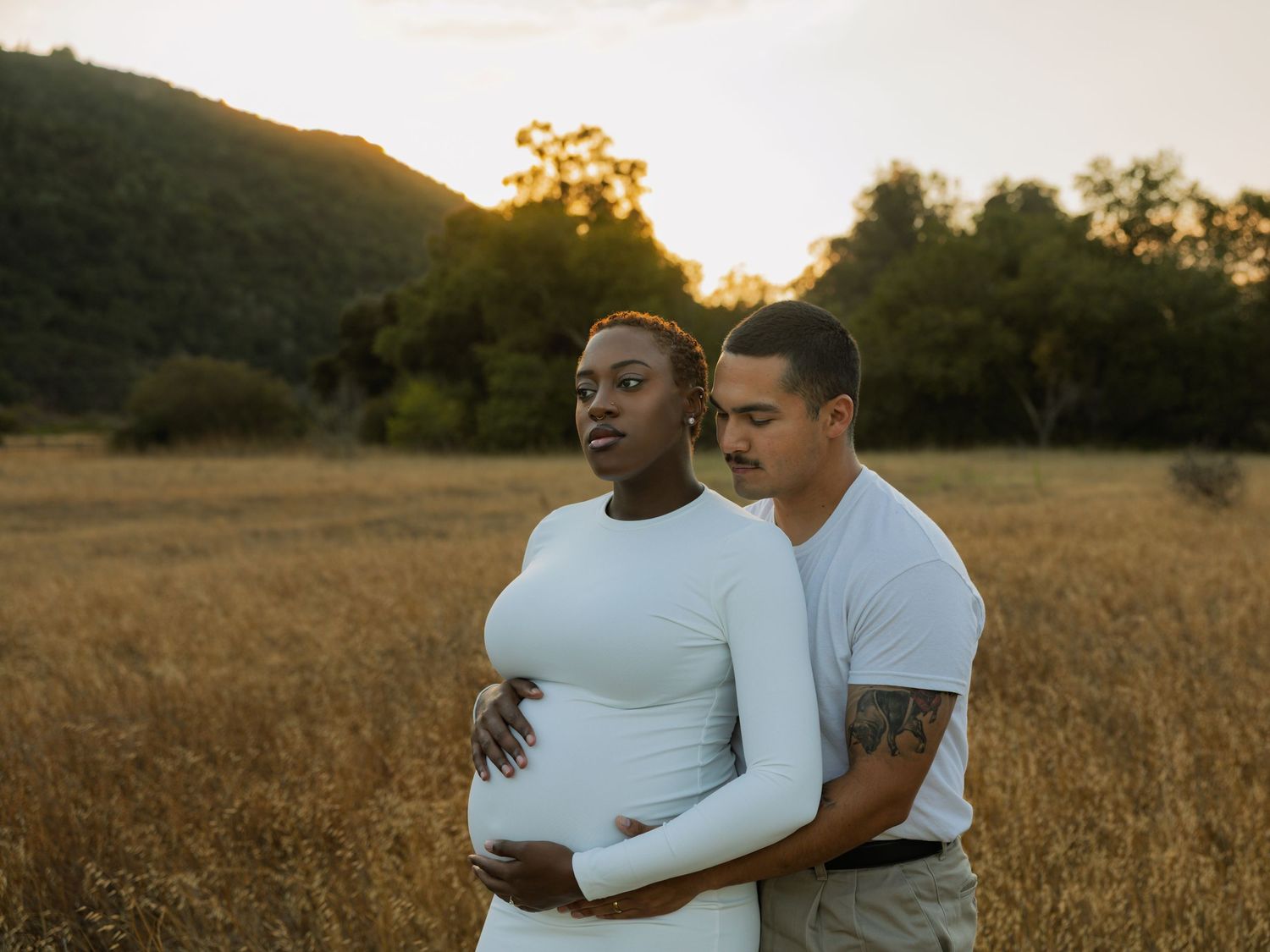 Pregnant couple embracing in a golden field at sunset, man holding woman's baby bump tenderly.