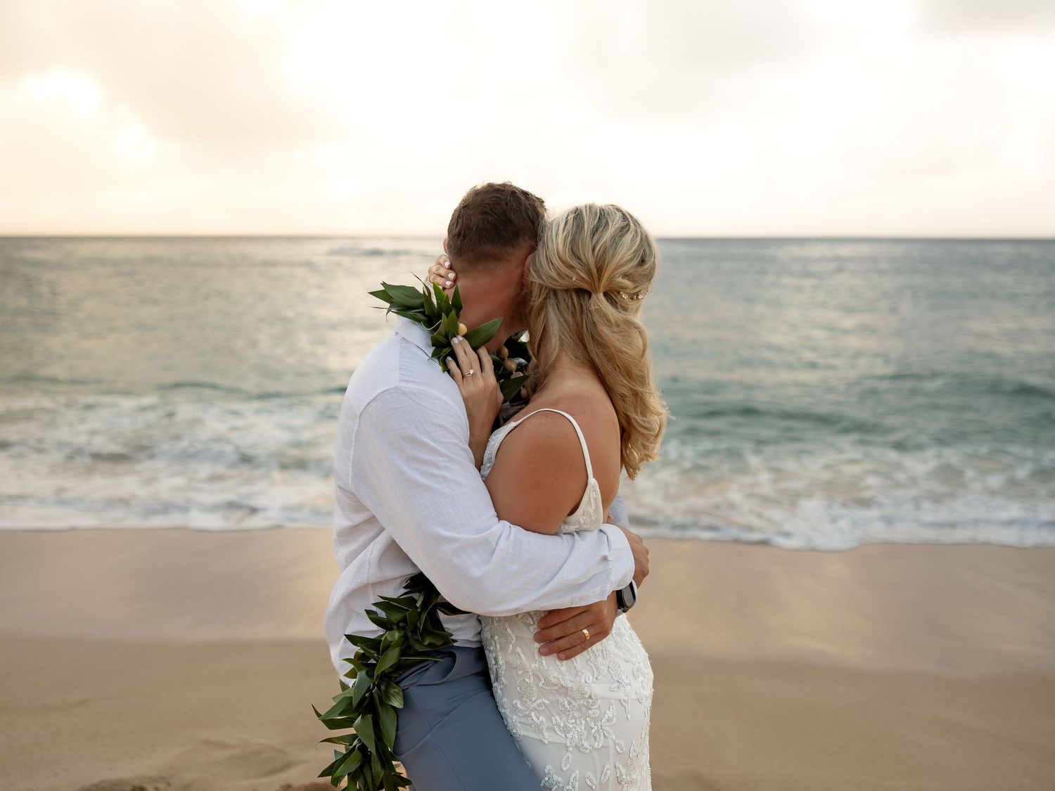 Couple sharing a romantic kiss on a sandy beach at sunset, bride in lace gown, groom with floral lei.