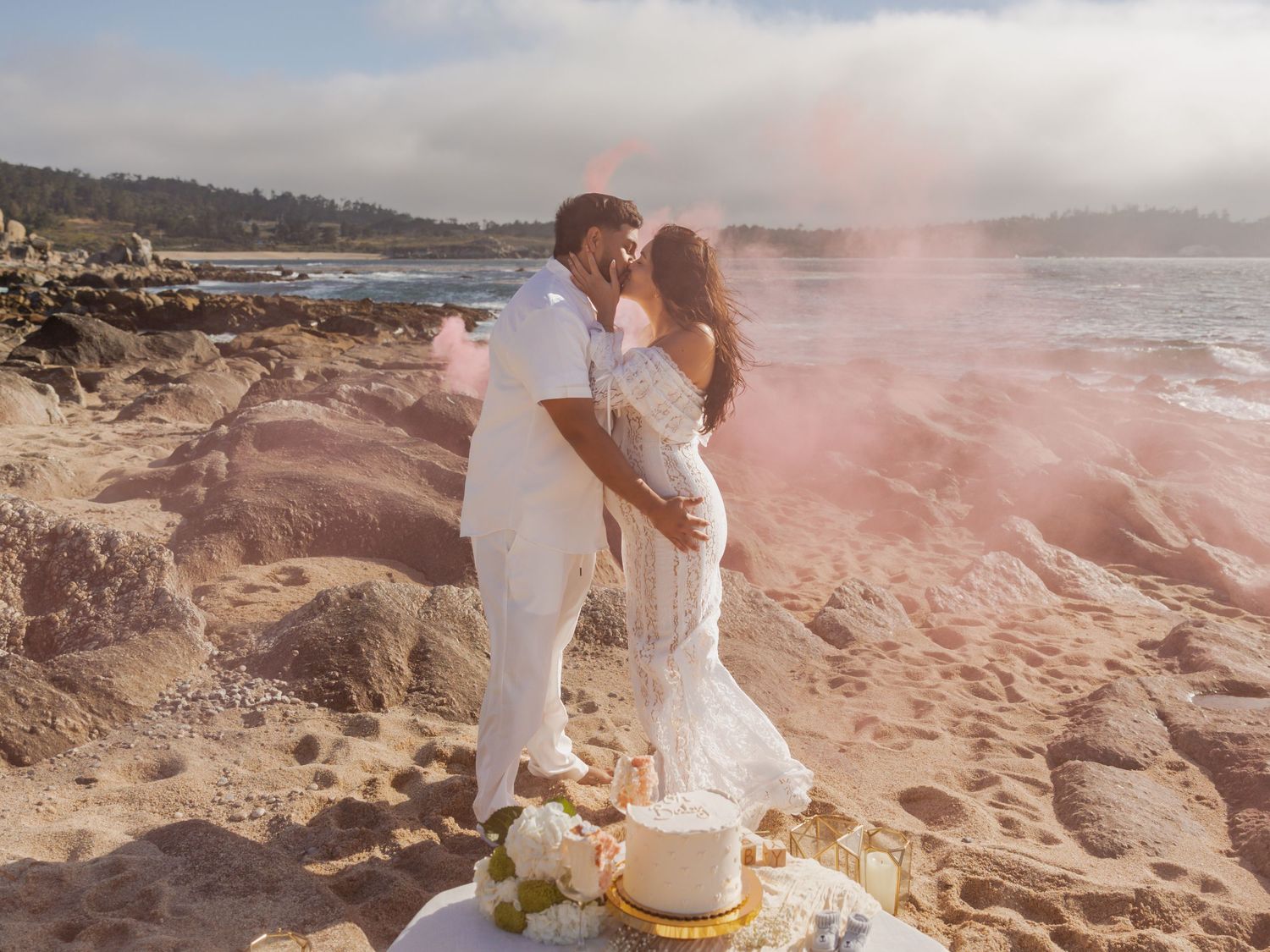 Couple kisses on rocky beach with pink smoke and a white cake, celebrating a gender reveal.