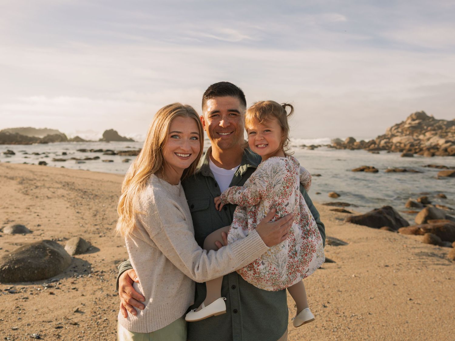 Happy family of three smiling together on rocky beach at sunset, parents holding young daughter.
