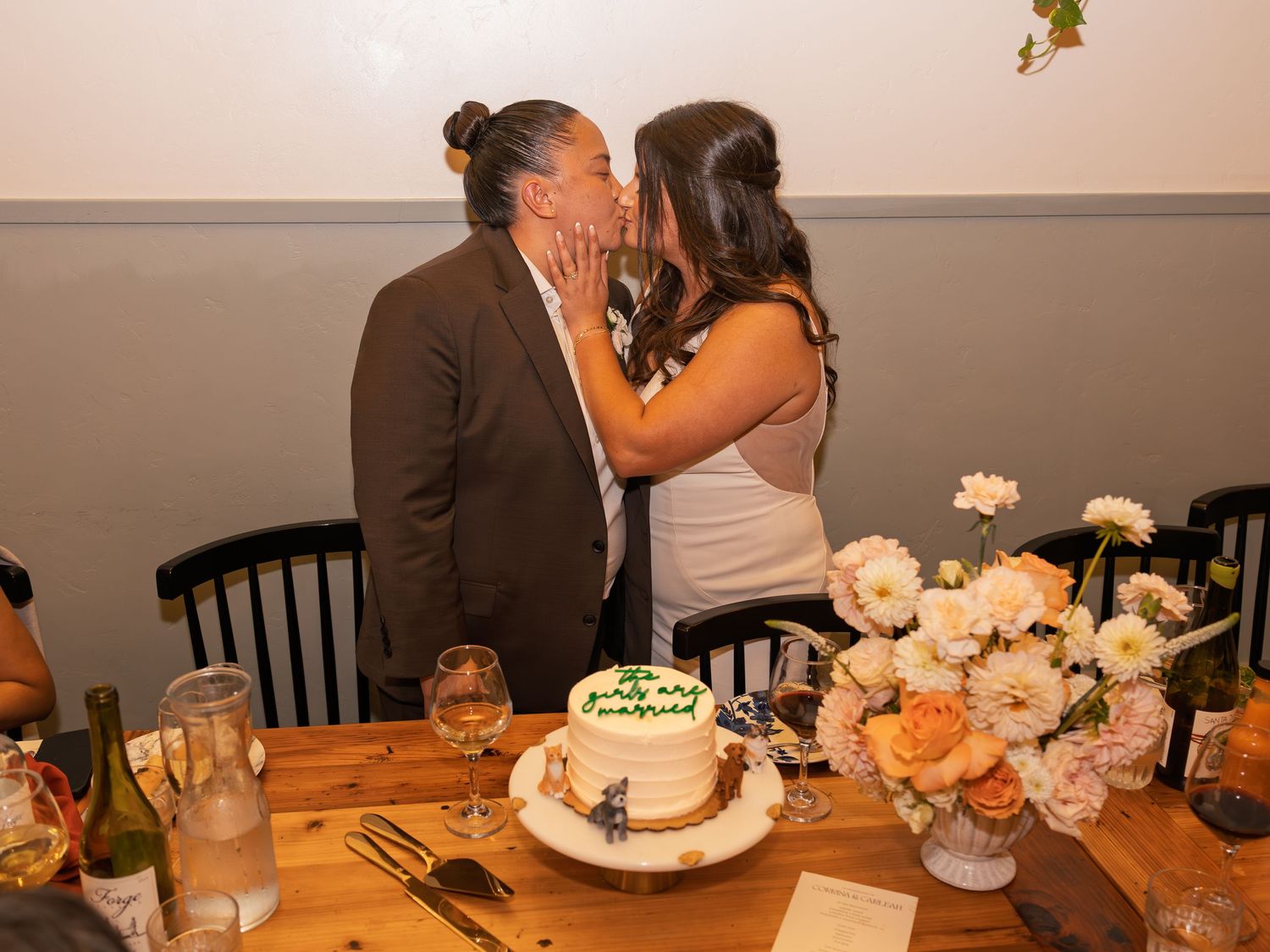 Couple sharing a kiss at wedding reception table with white cake and floral arrangement.