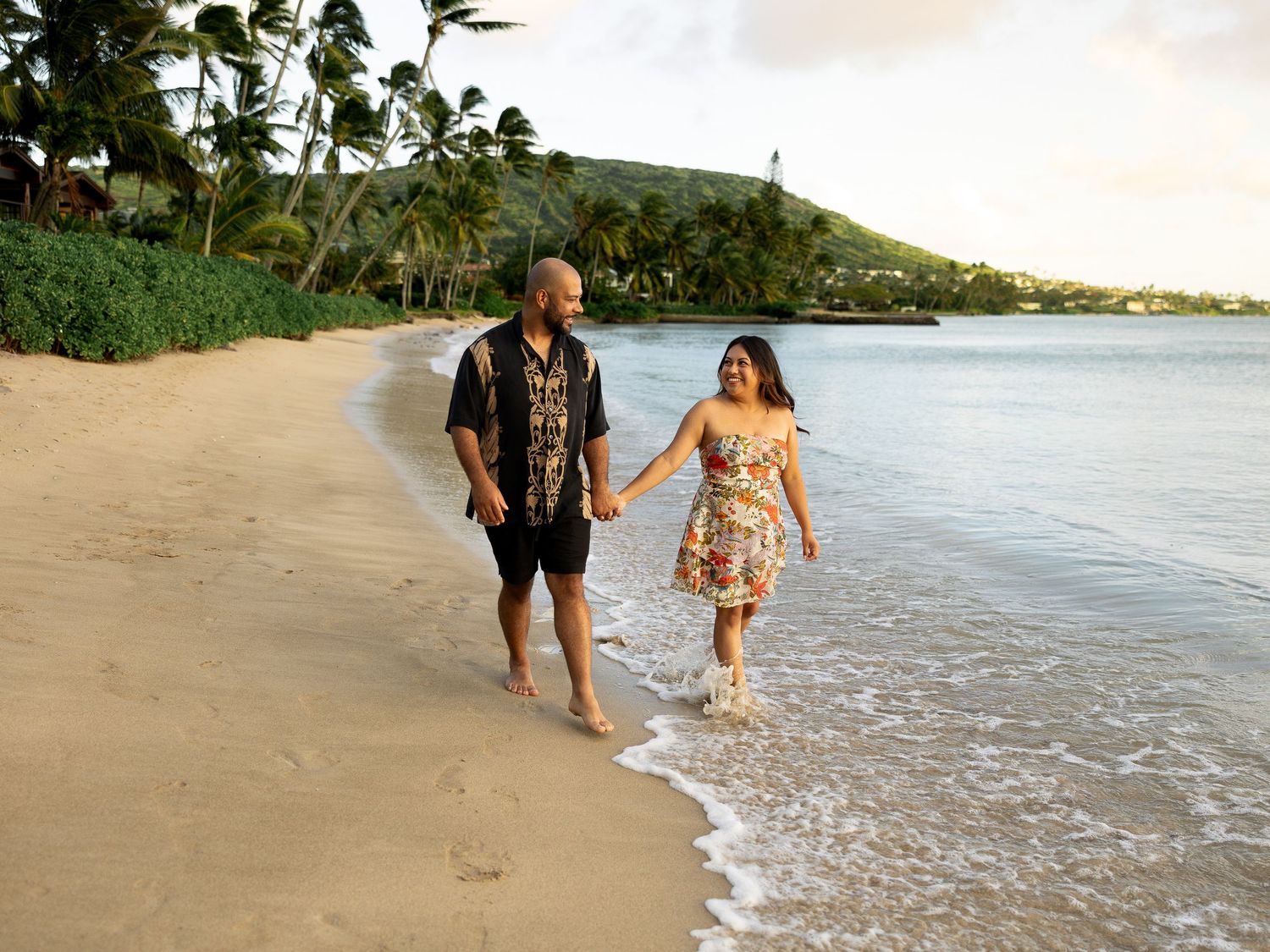 Couple holding hands walking barefoot on a sandy Hawaiian beach at sunset with palm trees and mountains.