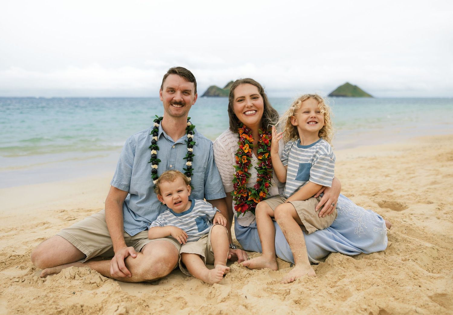 Family of four sitting on sandy beach in Hawaii, parents wearing leis, smiling with two young children.