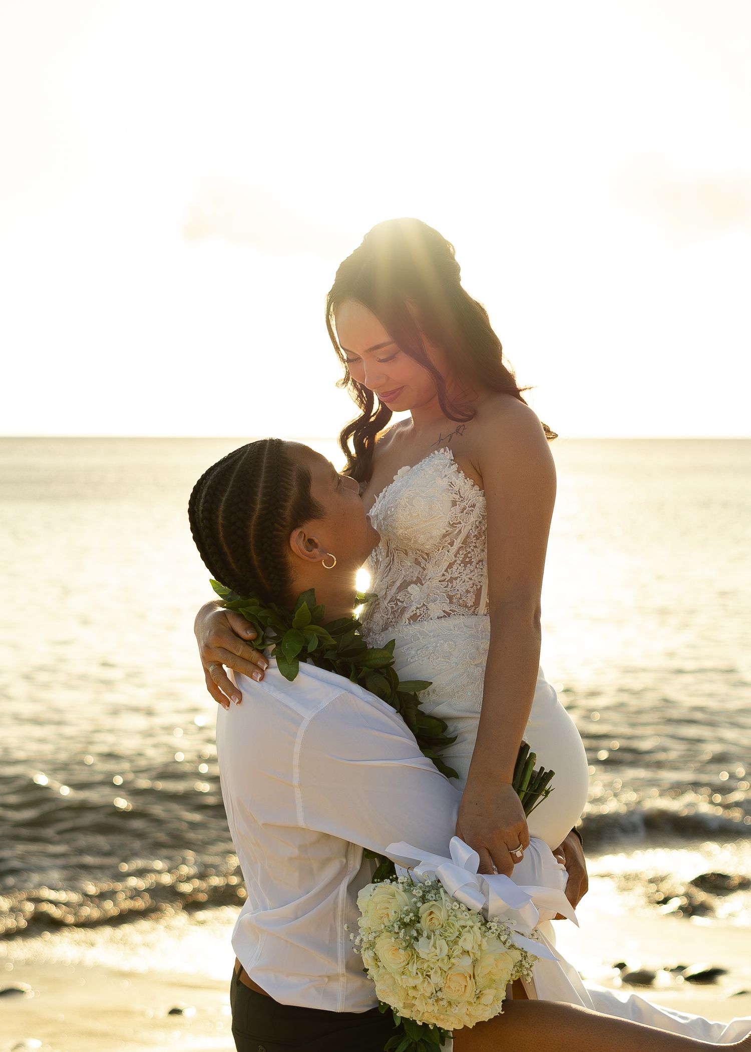 Couple sharing a romantic beach wedding moment at sunset, bride in lace gown holding white bouquet, groom in white shirt.