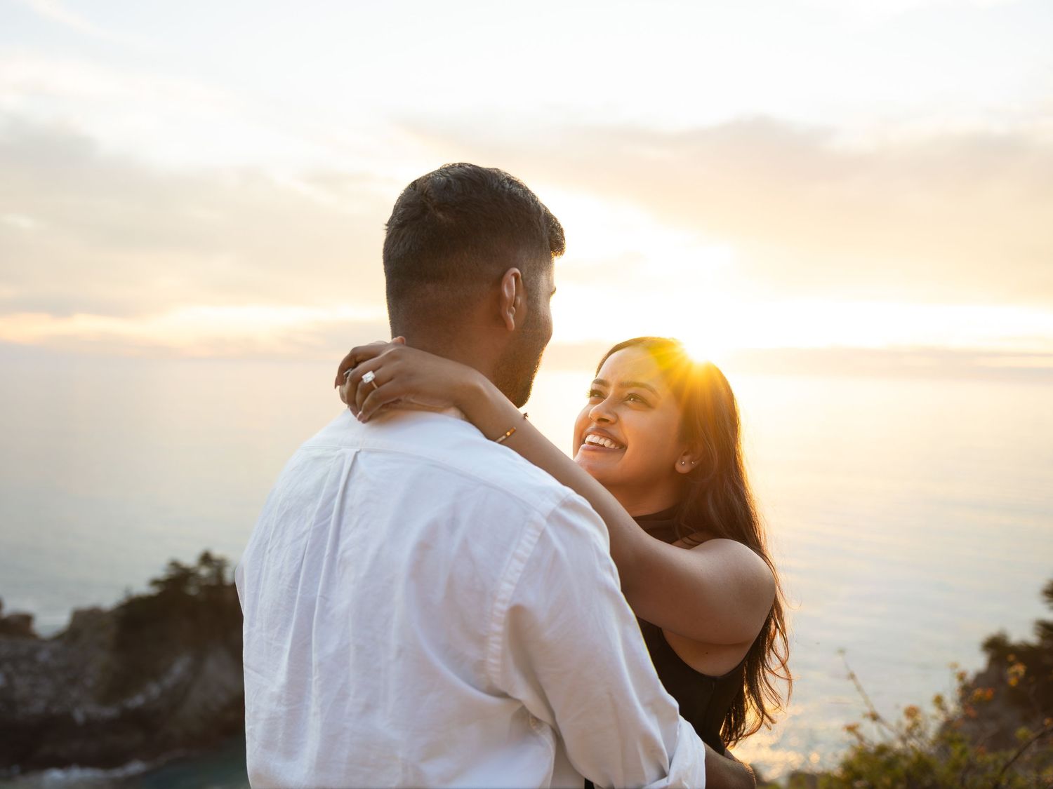 A couple embraces at sunset overlooking the ocean, the woman smiling warmly as golden light glows behind them.