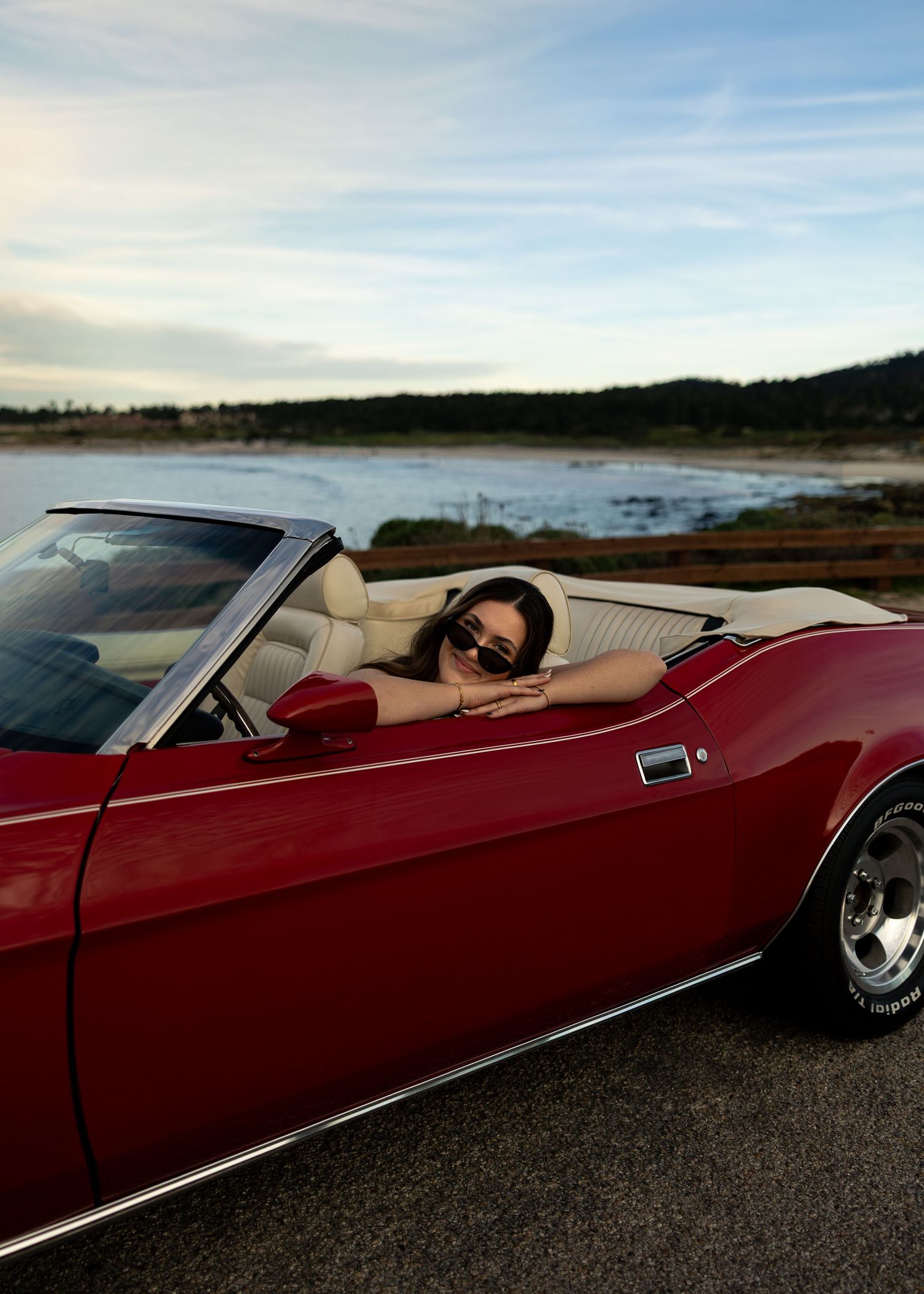 Woman in sunglasses leaning on a classic red convertible muscle car parked near a scenic waterfront at dusk.