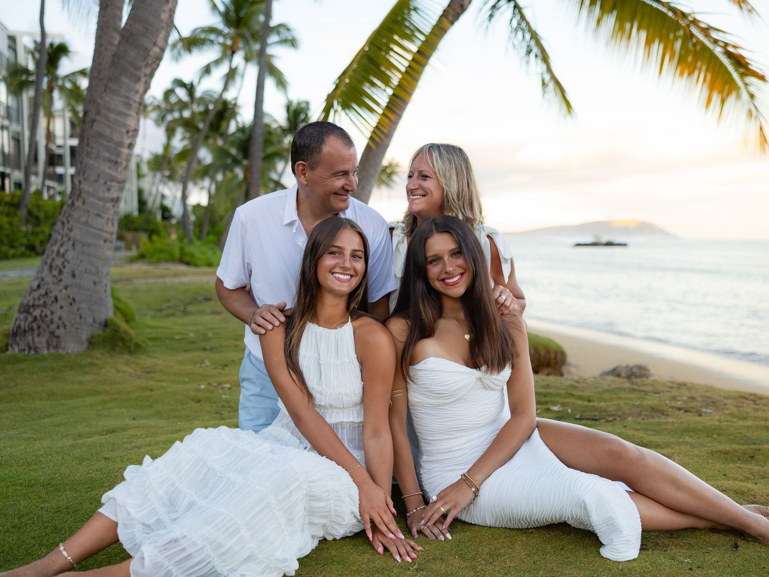 Family of four posing on tropical beach at sunset, wearing white outfits, smiling with palm trees and ocean in background.