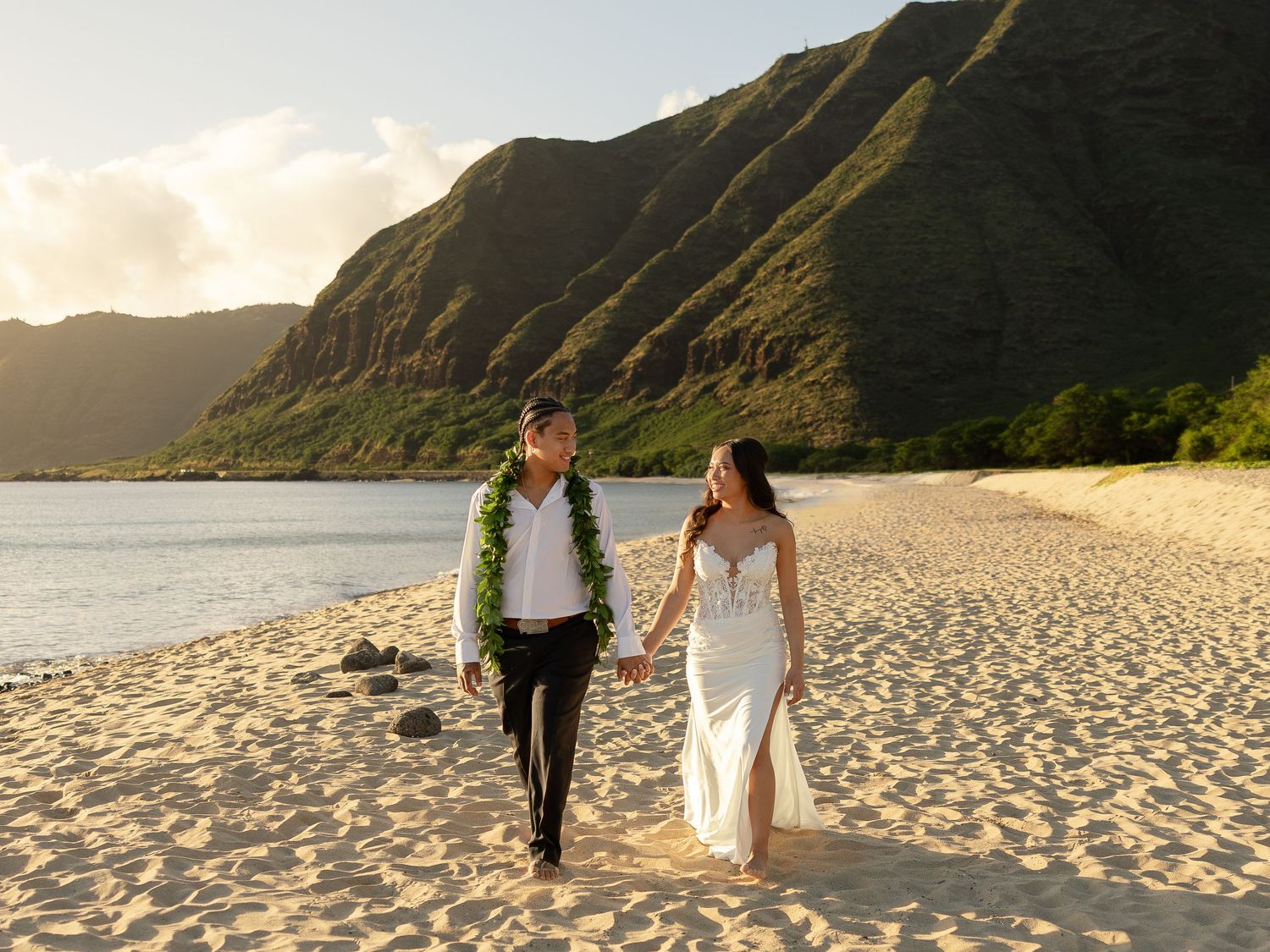 Newlywed couple walking hand in hand on a sandy Hawaiian beach with lush green mountains at sunset.