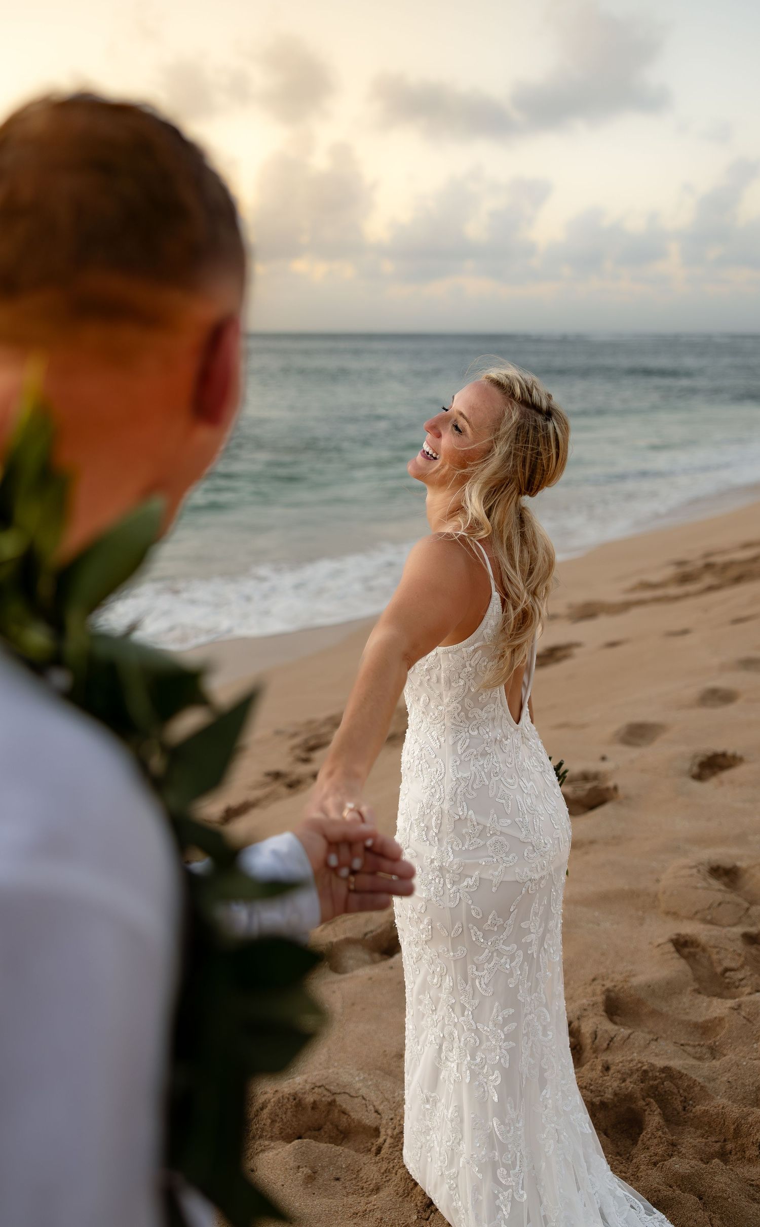 Bride in white lace gown holds groom's hand on sandy beach at sunset, laughing joyfully near ocean waves.