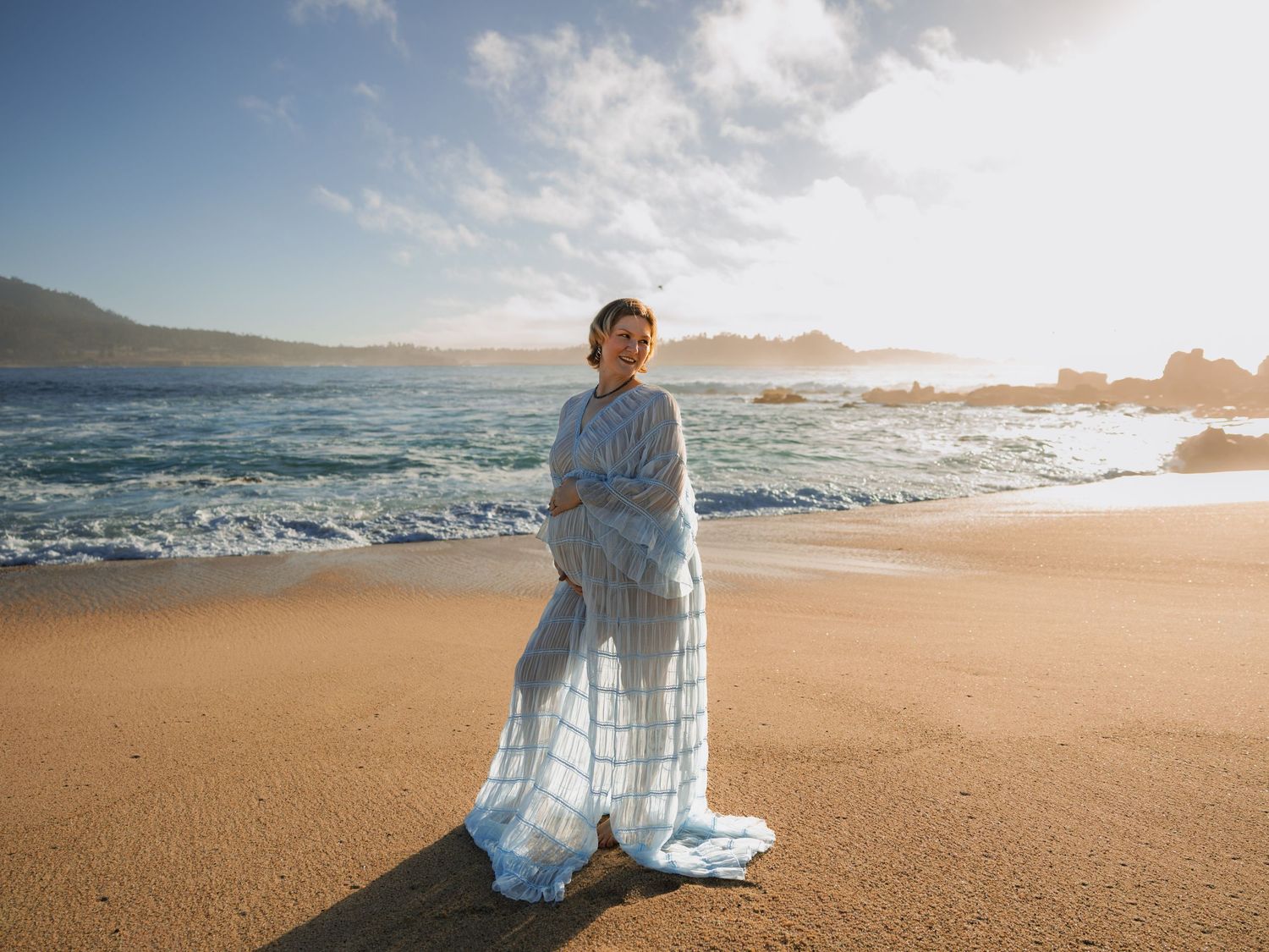 Pregnant woman in flowing blue dress standing on sandy beach with ocean waves and rocky coastline in background.