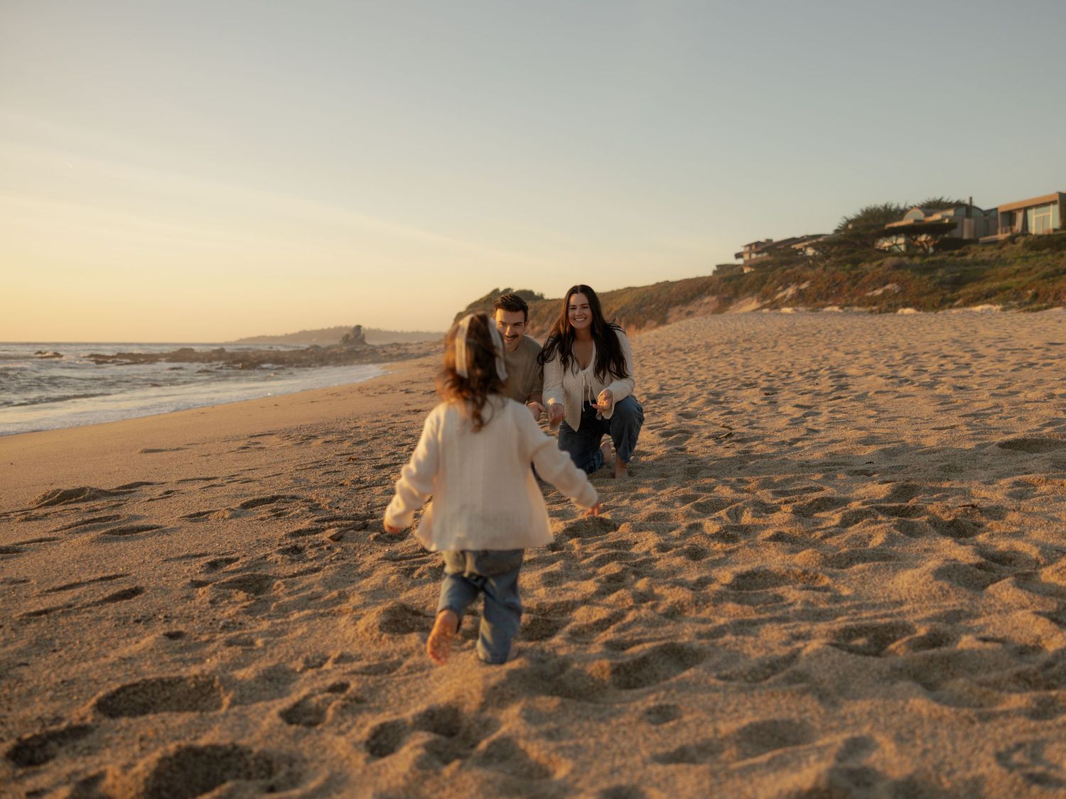 Family enjoying a sunset beach walk on golden sand with ocean waves and coastal homes in the background.