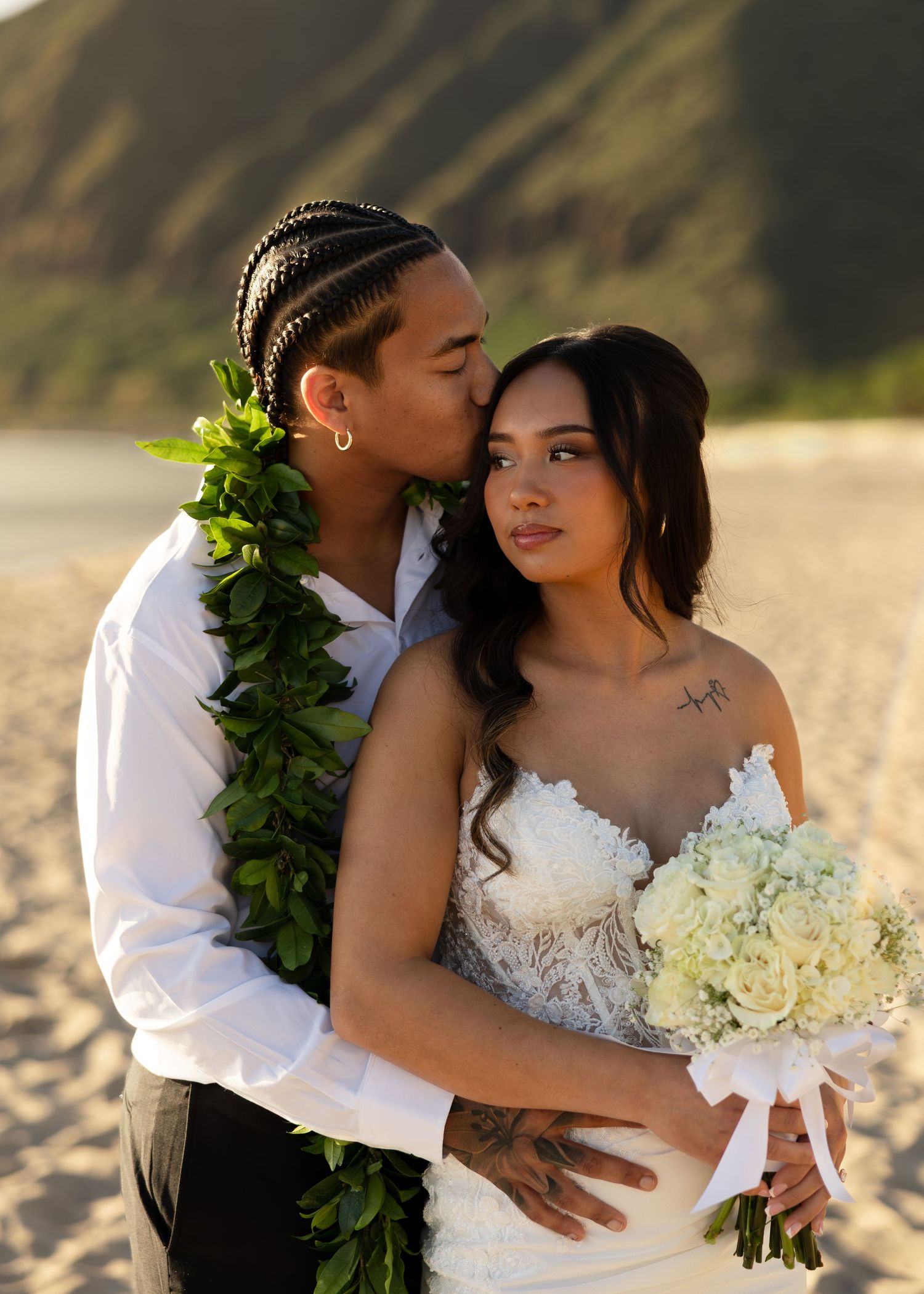 Groom in white shirt and green lei kisses bride in lace gown holding white bouquet on Hawaiian beach.