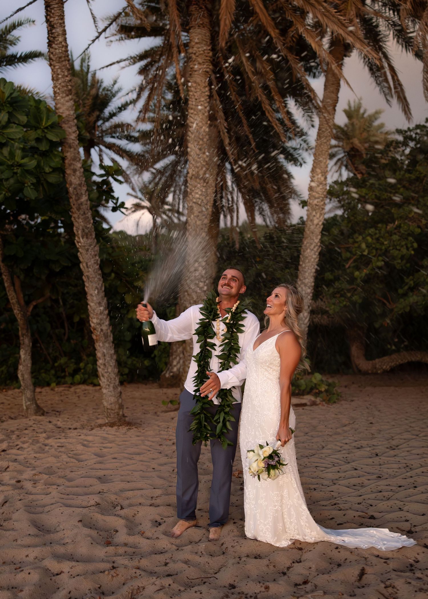 Newlywed couple celebrating on a tropical beach, popping champagne in wedding attire with palm trees backdrop.