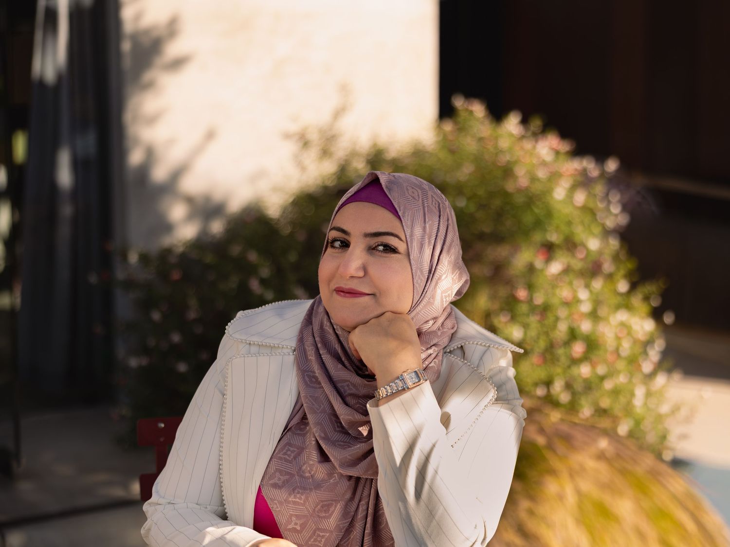 Woman in pink hijab and white jacket sitting outdoors, resting chin on hand with flowers in background.