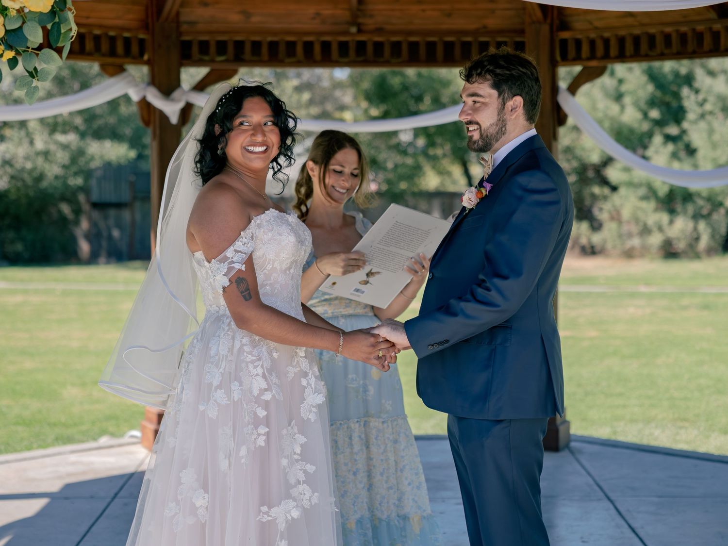 Smiling bride and groom exchange vows under a wooden gazebo outdoors, officiated by a woman holding a decorative book.