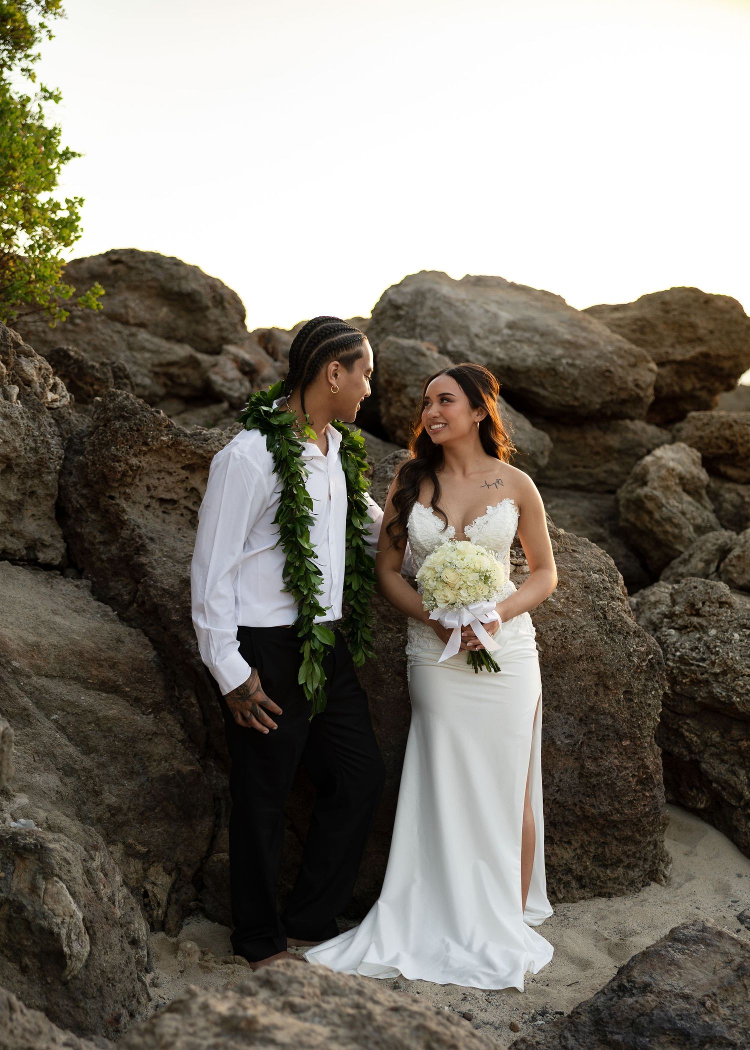Couple in wedding attire standing among rocks on a Hawaiian beach, groom wearing green lei, bride holding white bouquet.