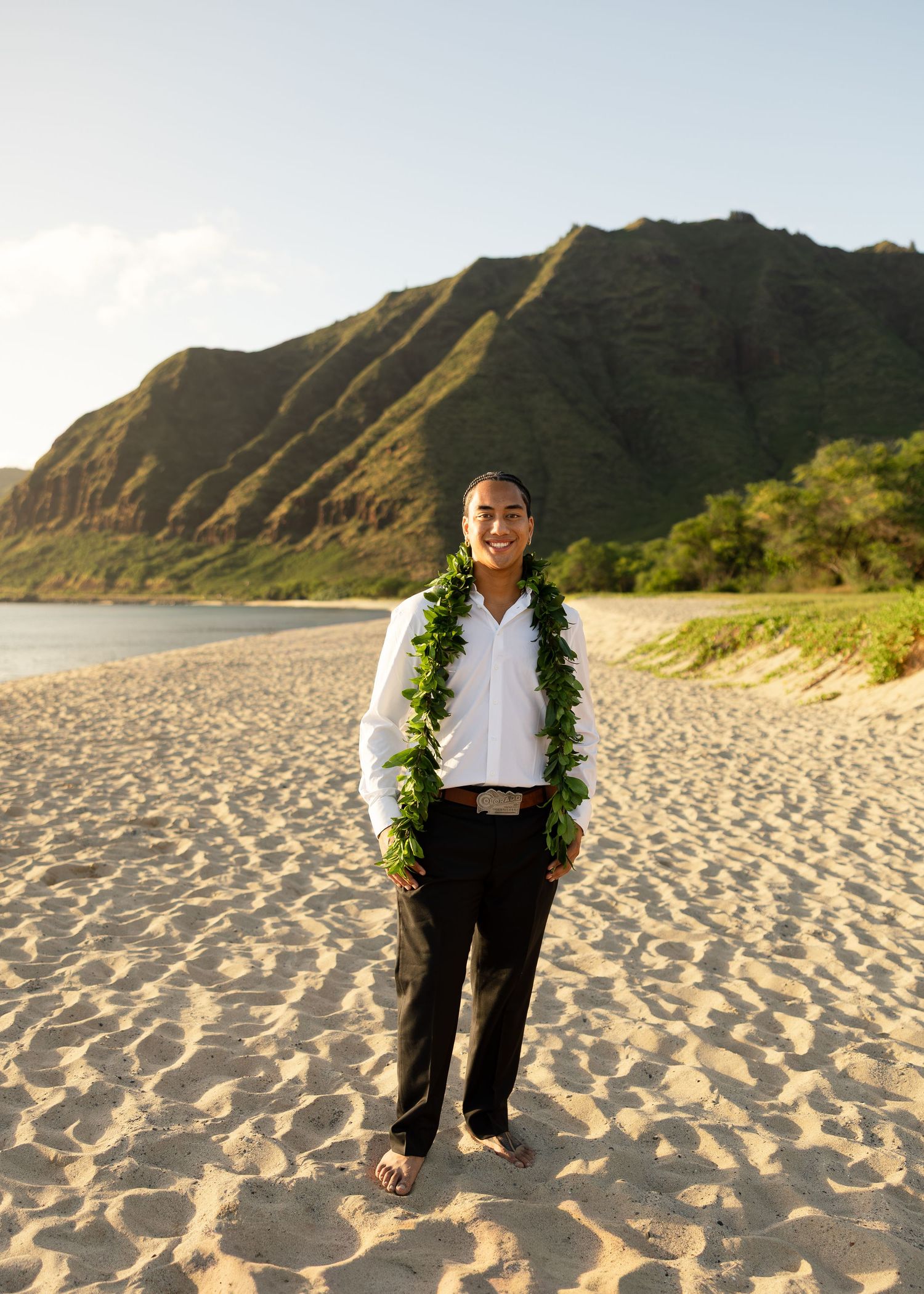 Young man wearing a green lei stands smiling on a Hawaiian beach with lush green mountains in the background.