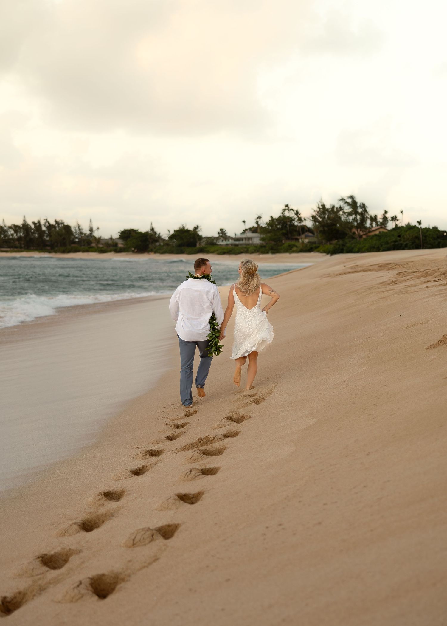 Couple walking hand-in-hand on a sandy beach, leaving footprints, with ocean waves and tropical trees in the background.