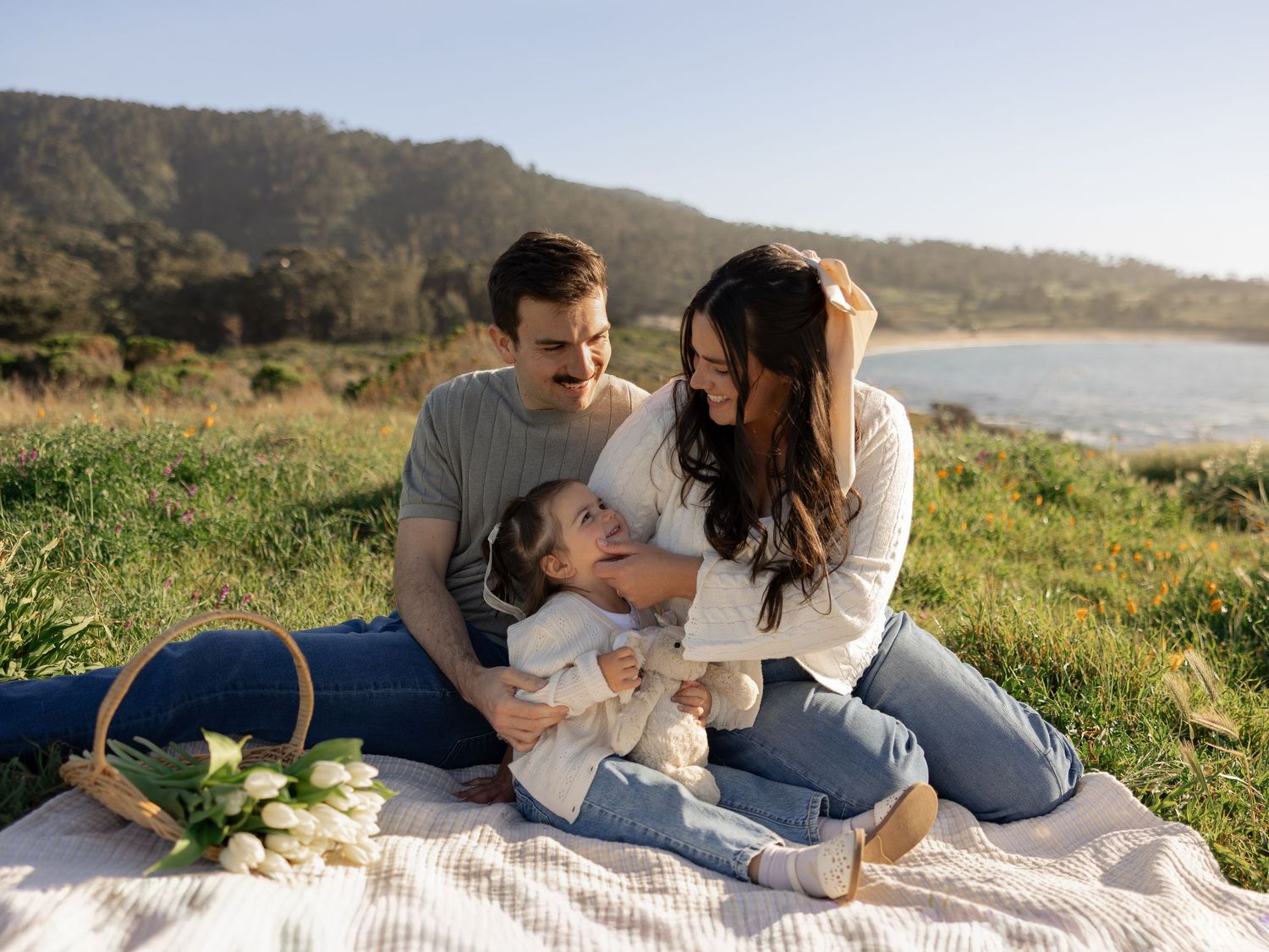 Happy young family with baby enjoying a sunny picnic on green coastal hillside with ocean views.