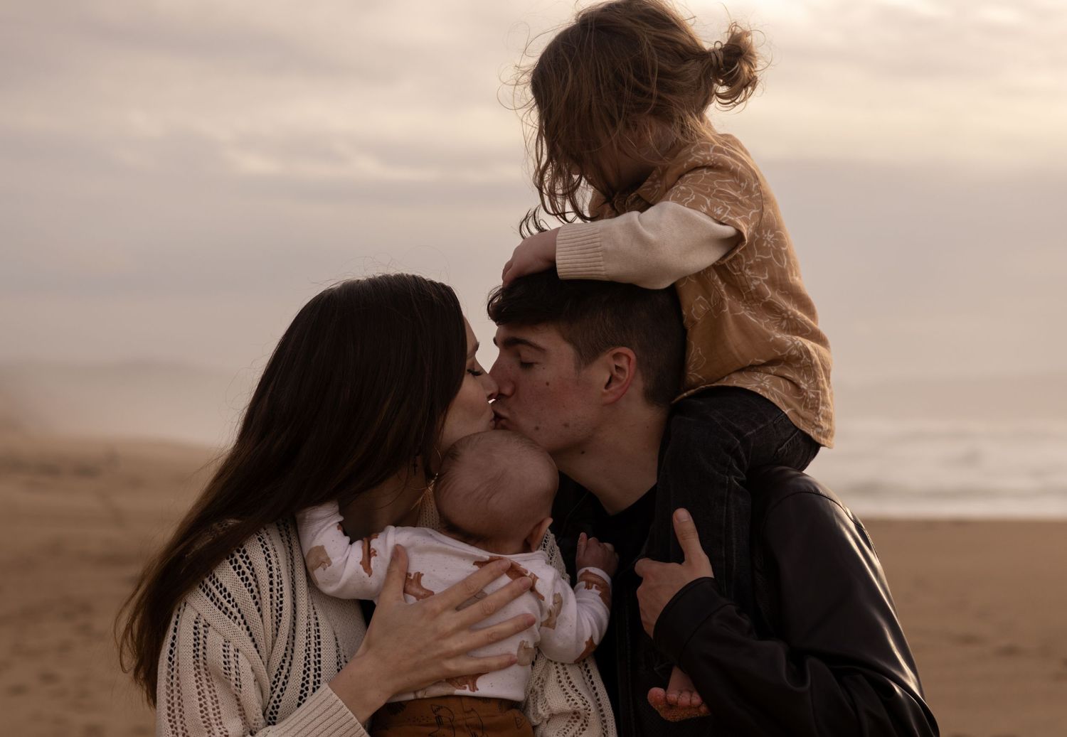 Family sharing a tender moment on the beach, parents kissing while holding baby and toddler on shoulders at sunset.