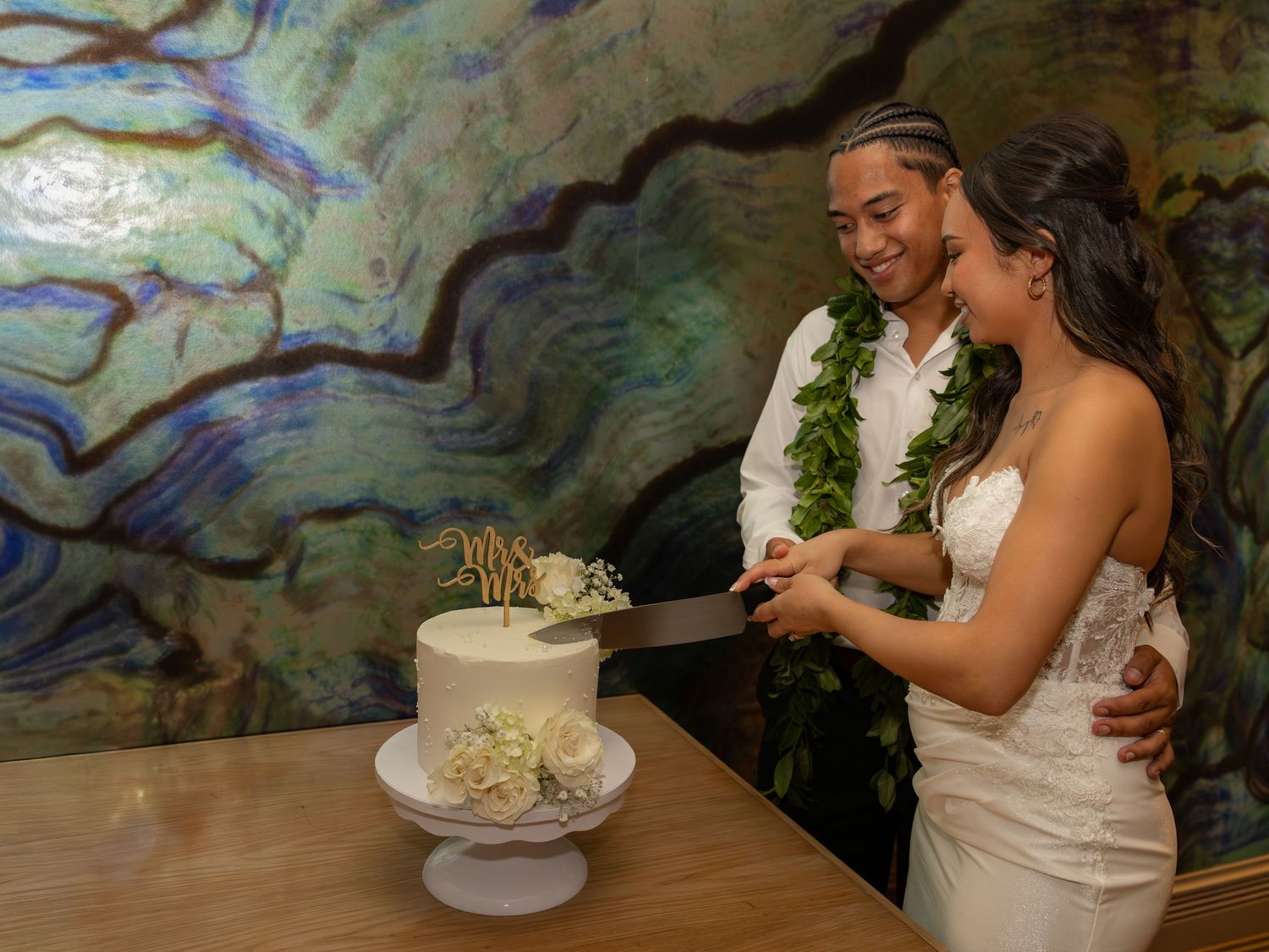 Newlywed couple cutting white wedding cake wearing floral leis, elegant venue with colorful marble wall backdrop.