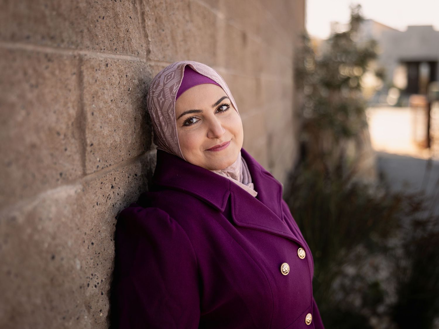Woman in purple hijab and coat leaning against stone wall, smiling outdoors.