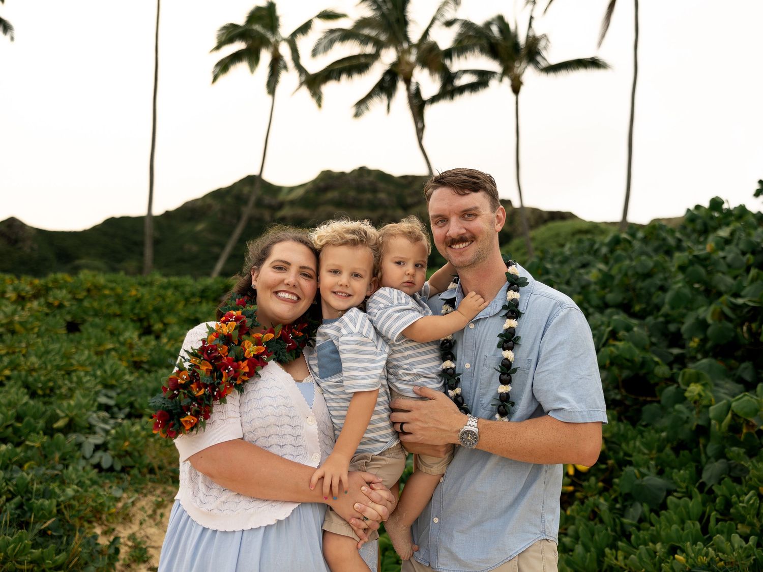 Family of four smiling outdoors in Hawaii, wearing leis, with tropical palm trees and green hills in the background.