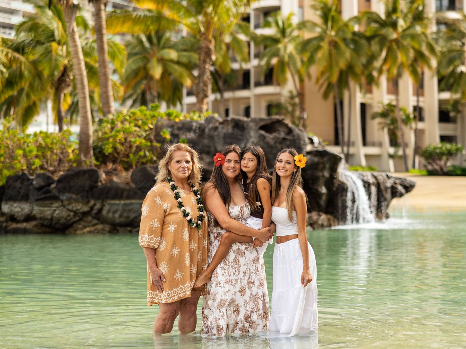 Four women posing together in a lagoon pool at a tropical Hawaiian resort with palm trees and waterfall.