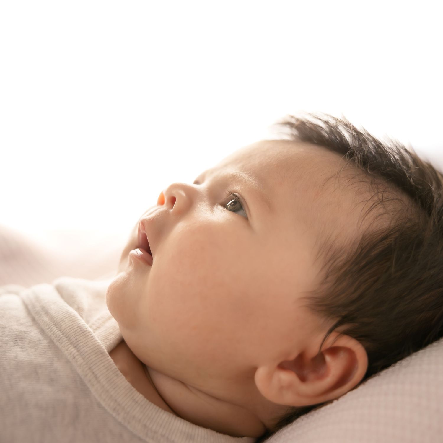 A peaceful newborn baby with dark hair lying down, gazing upward in soft natural light.